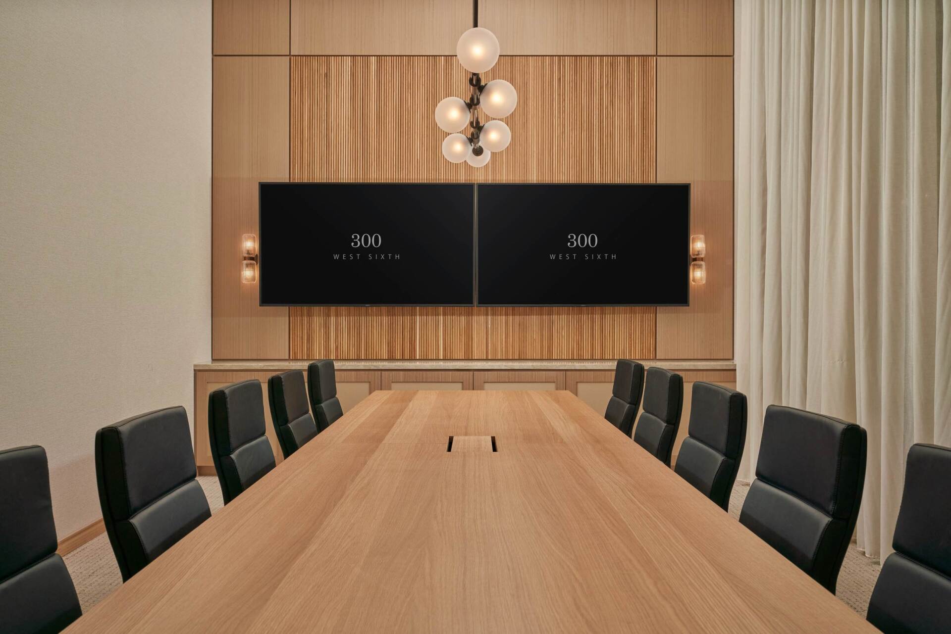 Conference room with long wooden table, black chairs, two screens, and light fixture on wood-paneled wall.