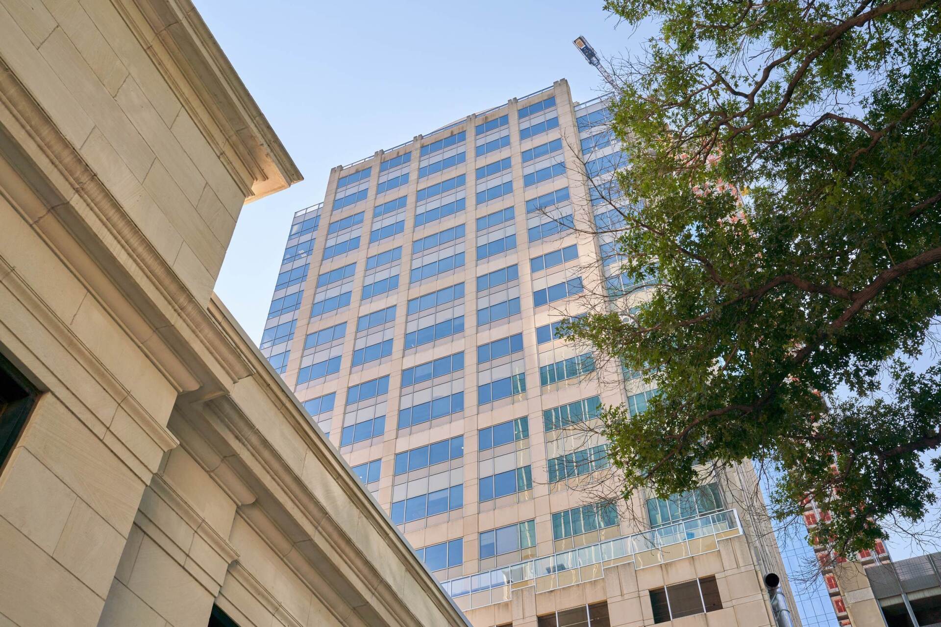 Tall building with many windows, seen from below, partially framed by a beige stone building and green tree branches.