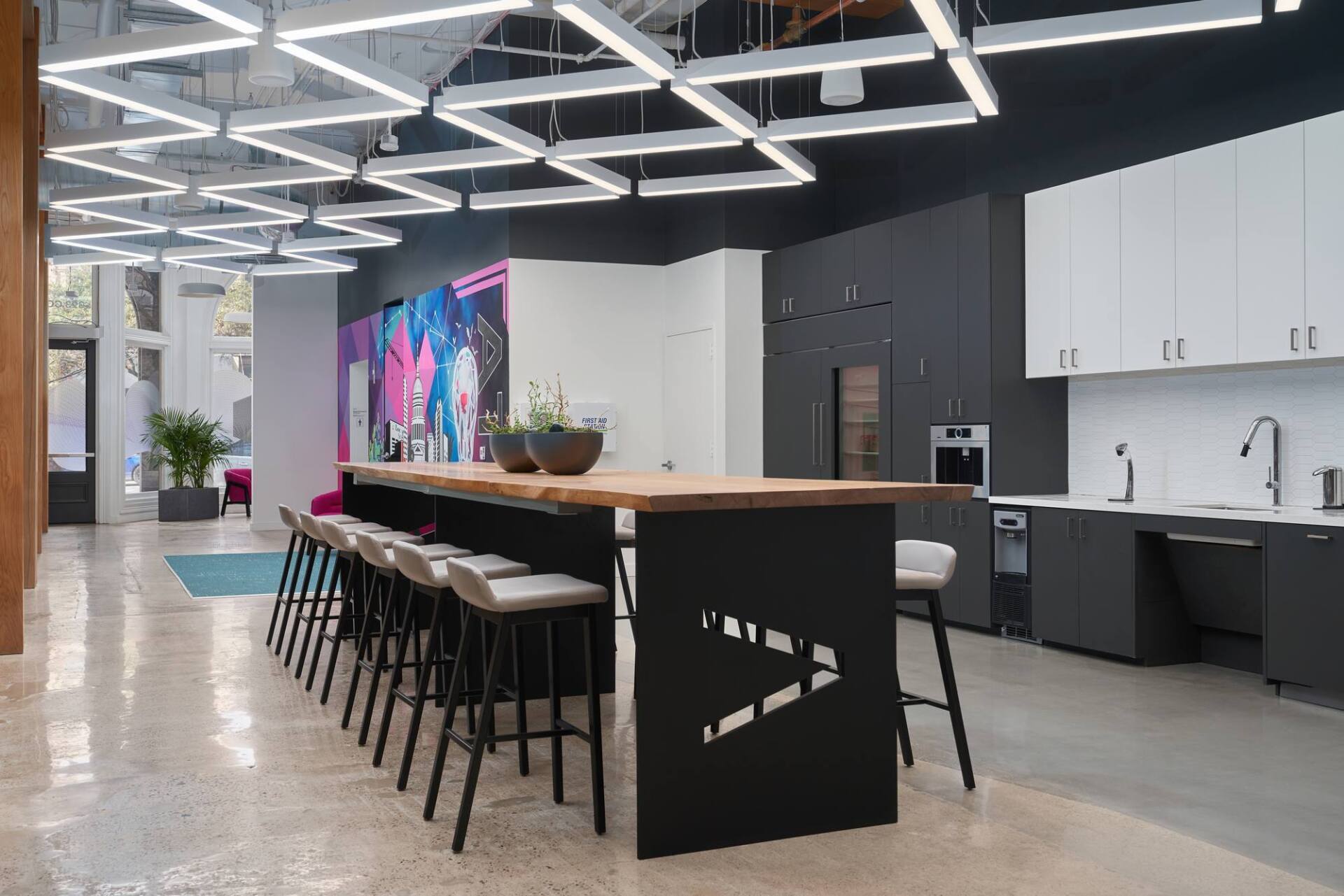 Modern office kitchen with a large island, stools, cabinetry, and geometric lighting.