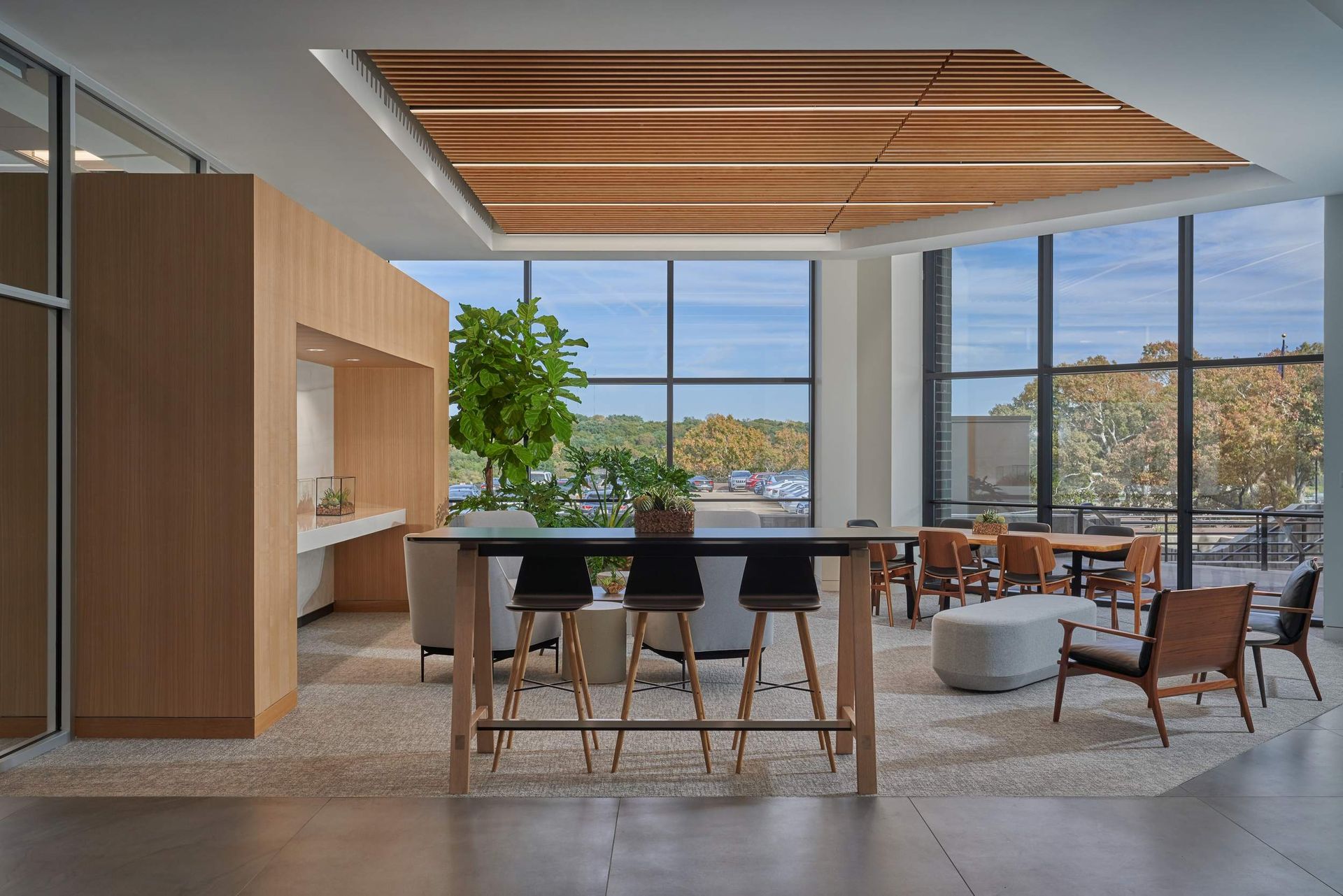 Modern office lounge with large windows, wooden accents, and a long table with stools.
