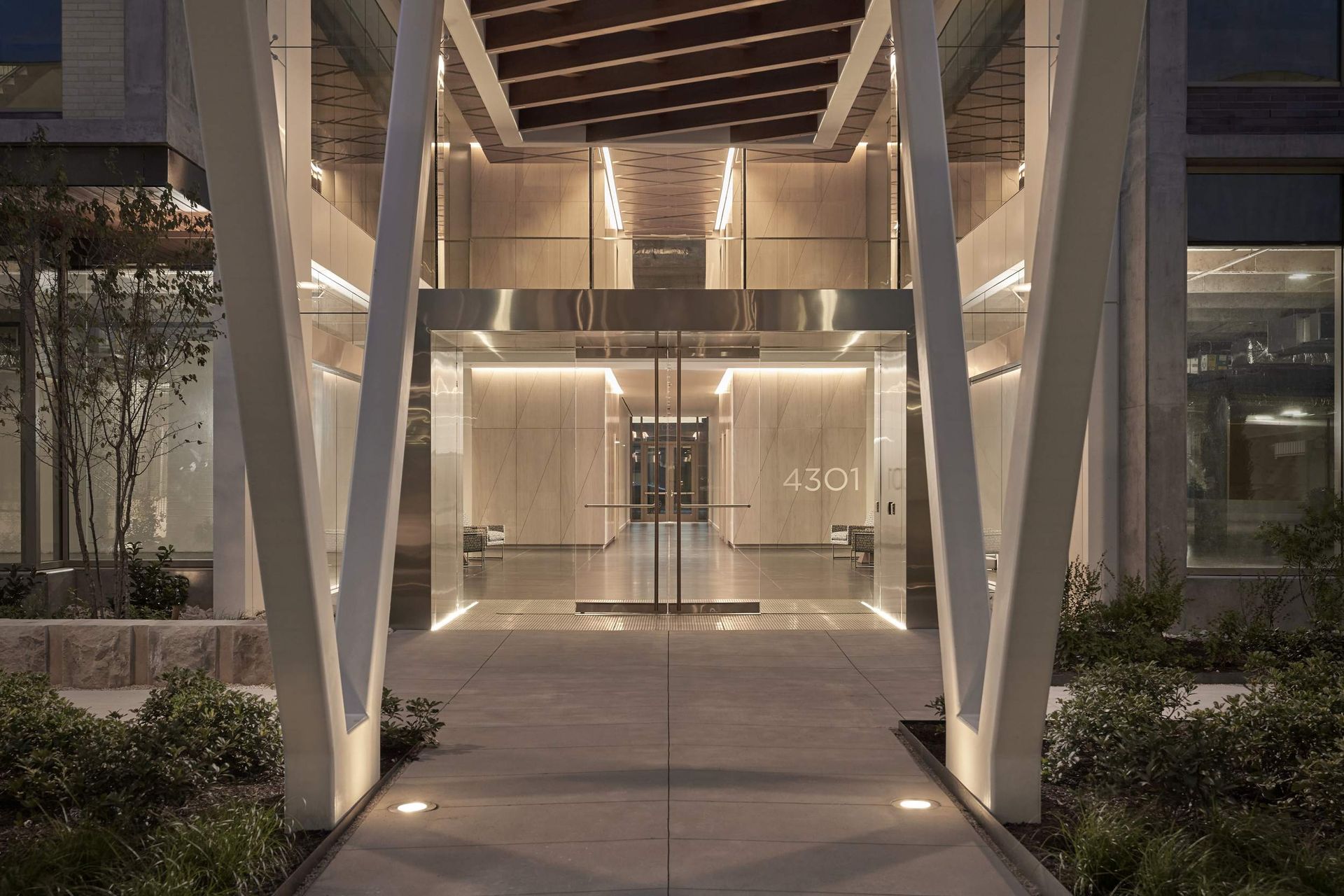 Entrance to a modern building at night, with illuminated walkways, glass doors, and architectural support beams.