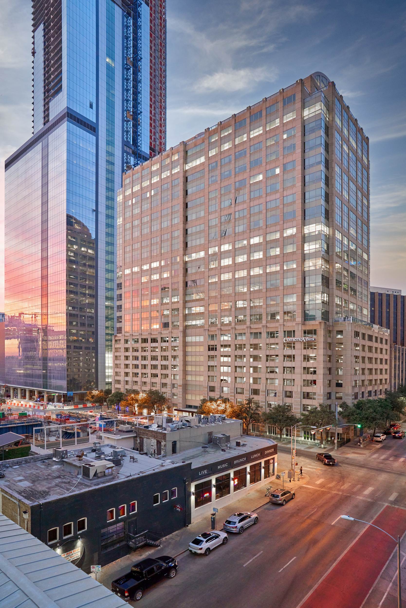 High-rise buildings at sunset: multi-story structures of glass and stone, street below, cars parked.