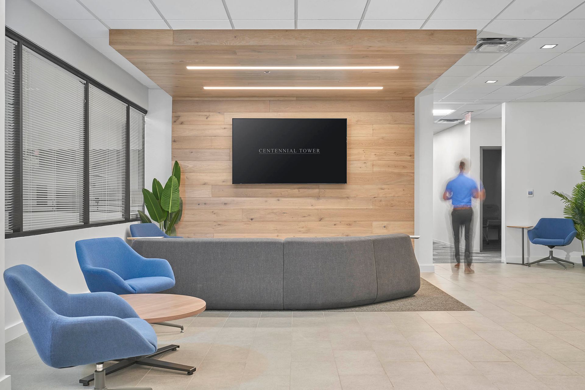 Modern office reception area with blue chairs, wood paneling, and a person walking in the background.