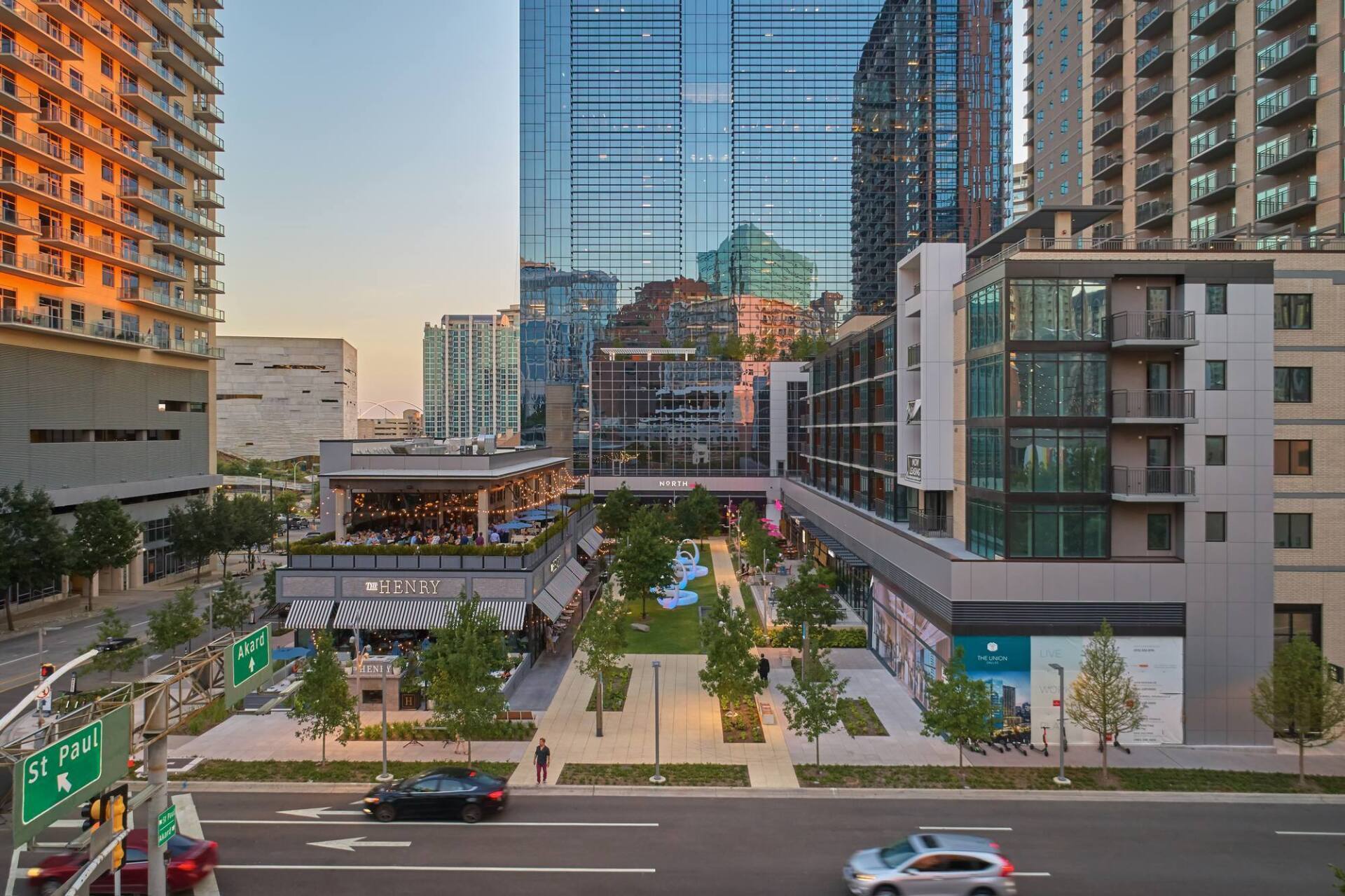 Urban street scene with buildings, park, and cars.