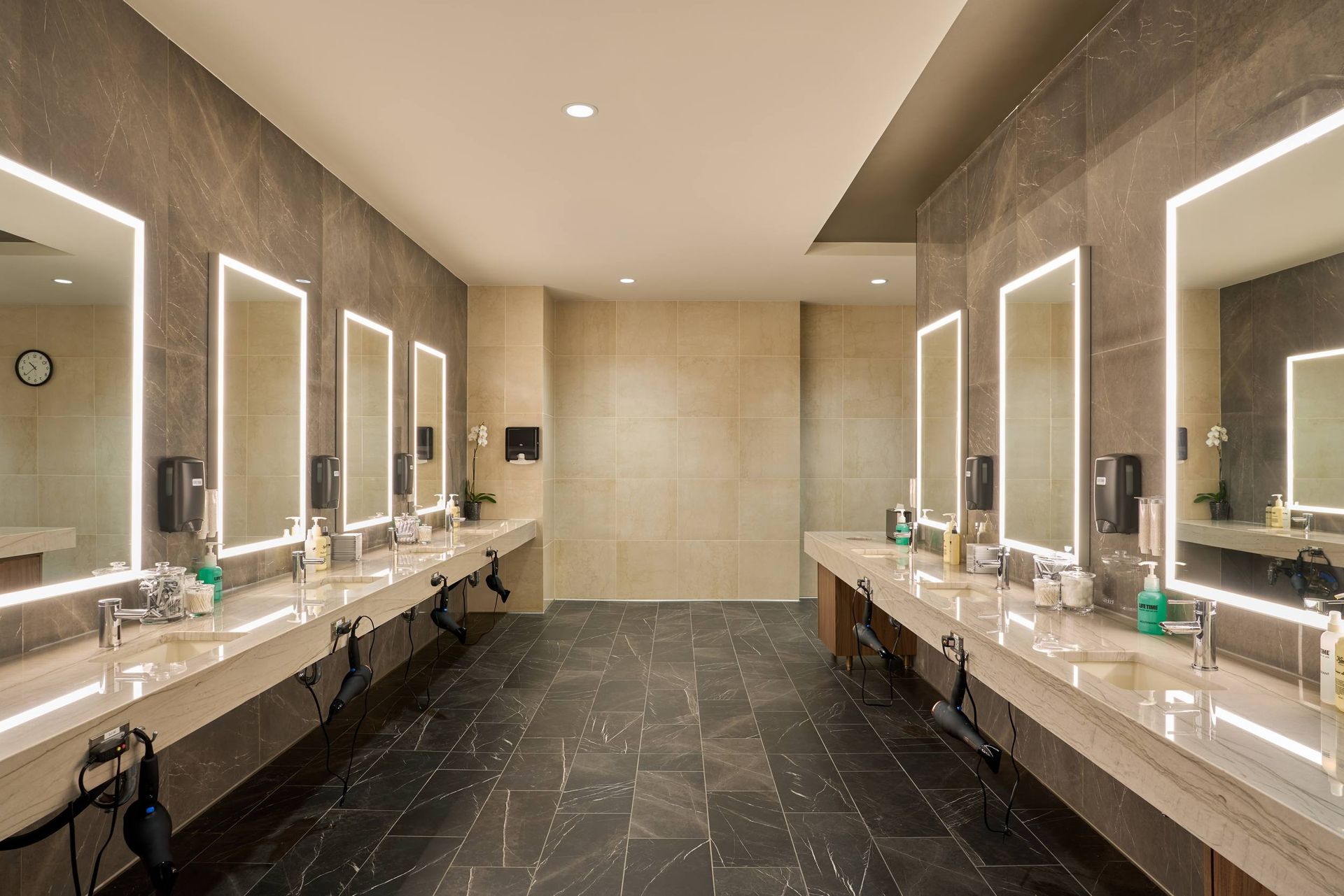 Spacious public restroom with multiple sinks, mirrors with LED lighting, and dark tile flooring.