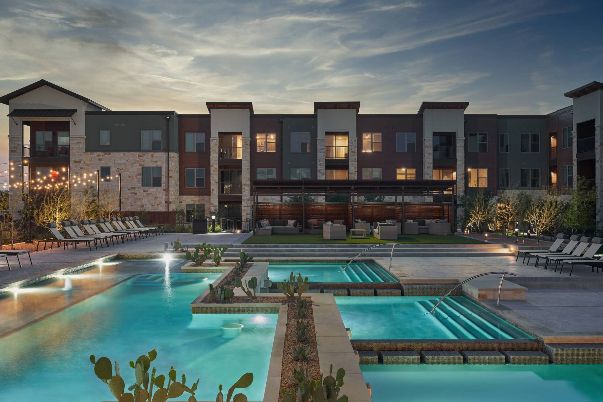 Apartment complex with a pool at dusk. Lit pool, lounge chairs, stone and stucco building, and string lights.