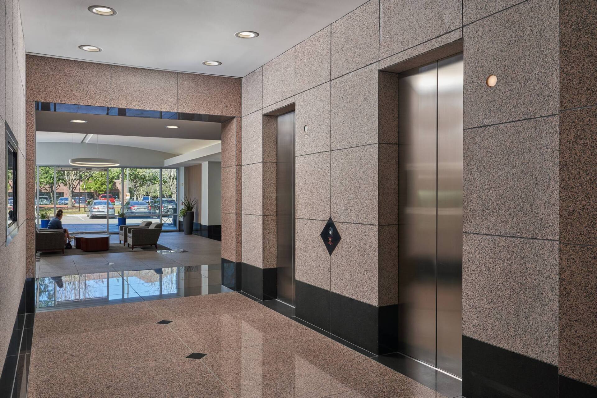 Lobby with two elevators, light-colored speckled walls and polished floor. View of seating area with windows.