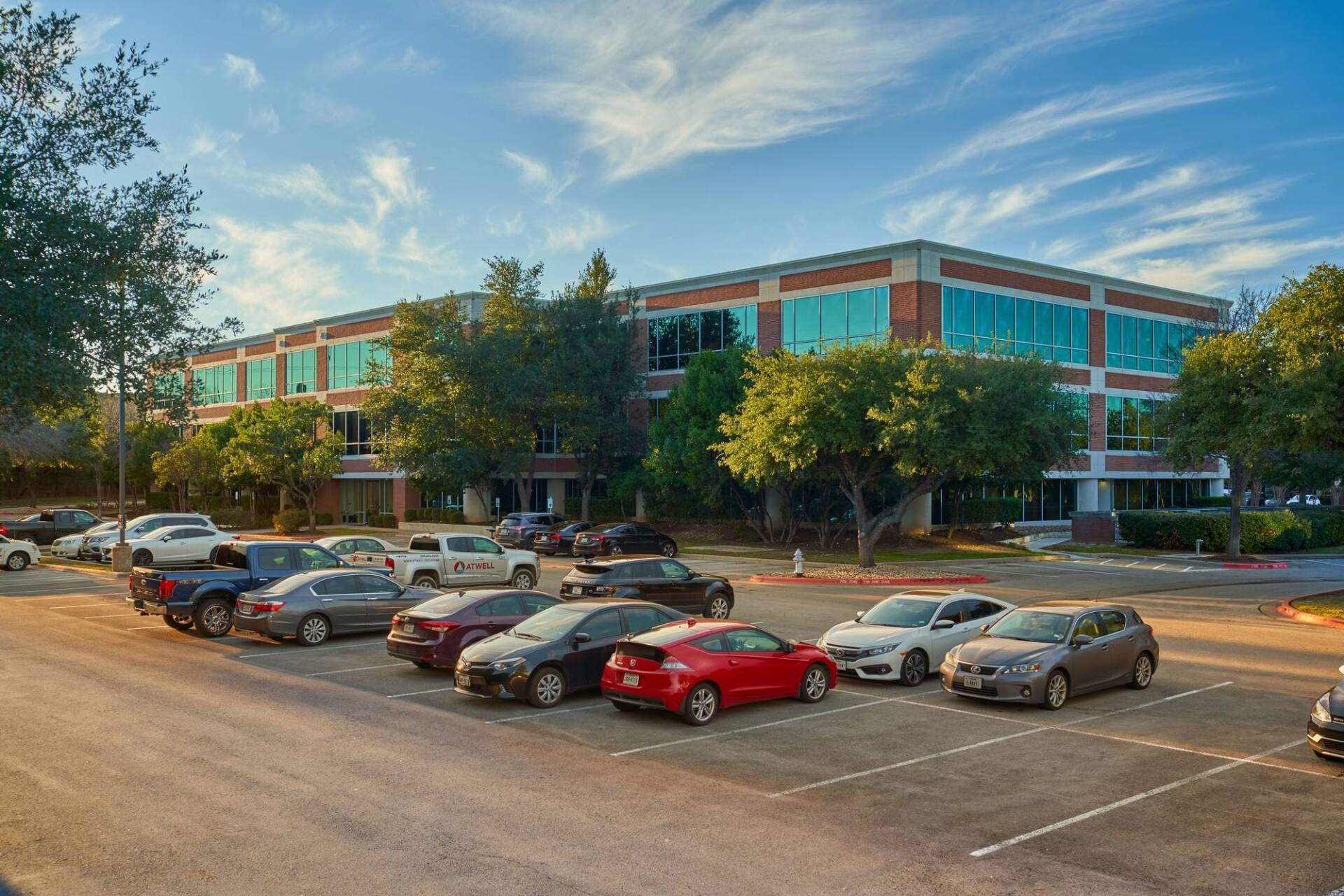 Office building with parked cars in a lot. Exterior view under a partly cloudy blue sky.
