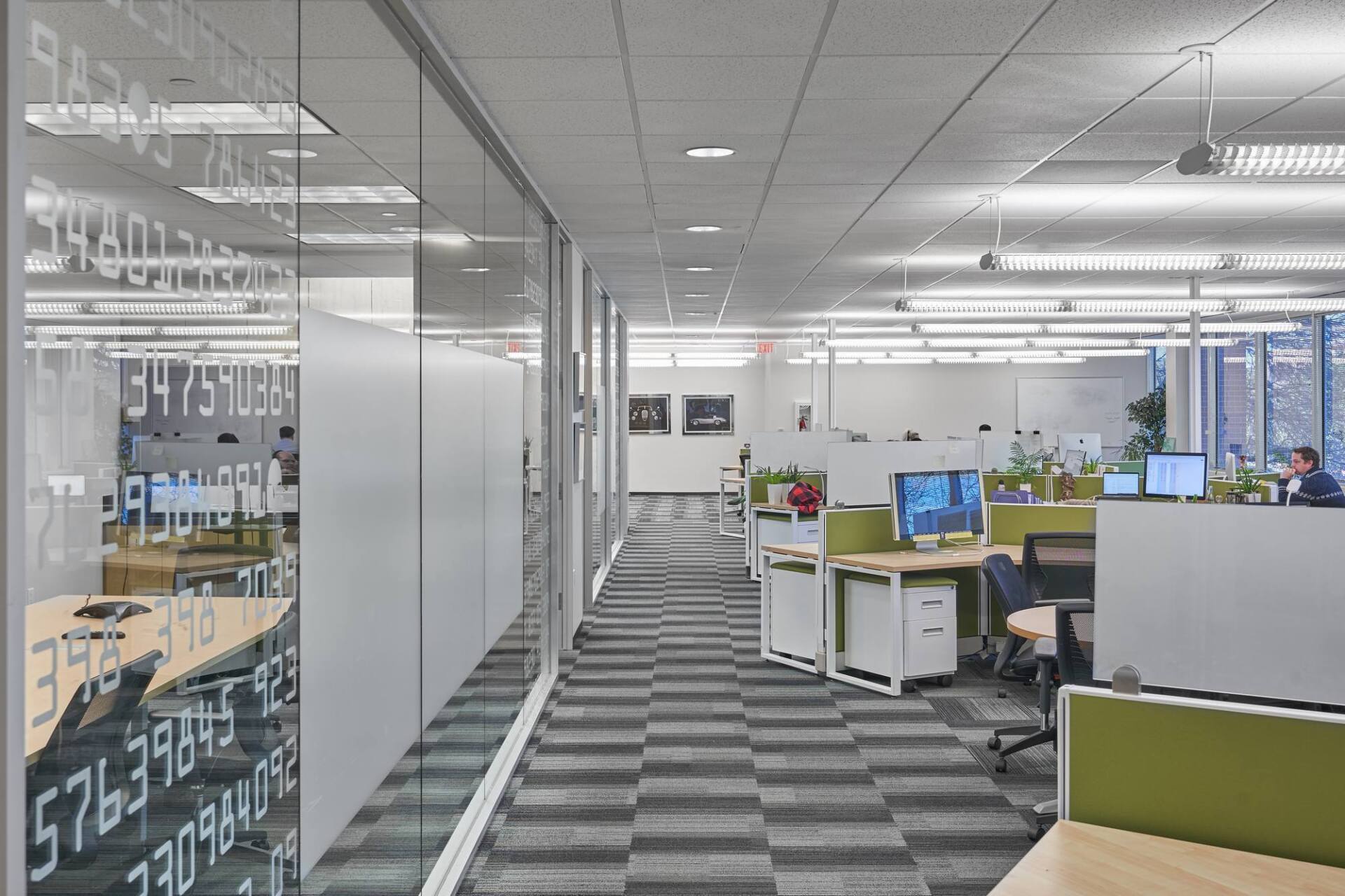 Office hallway with glass walls, cubicles, and a checkered carpet. A person works at a desk.