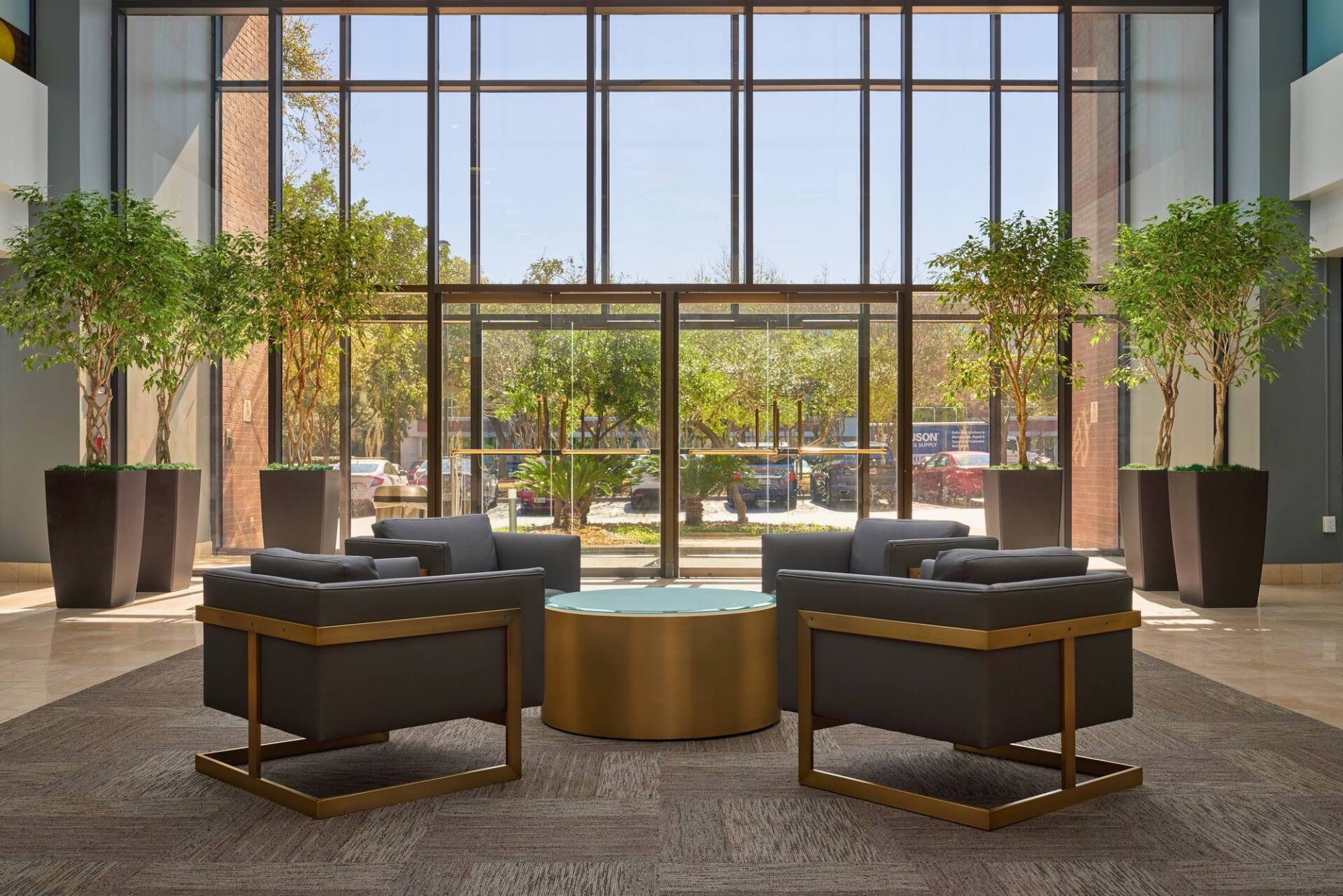 Lobby with armchairs, a gold table, and plants, in front of a glass wall with a view of greenery.