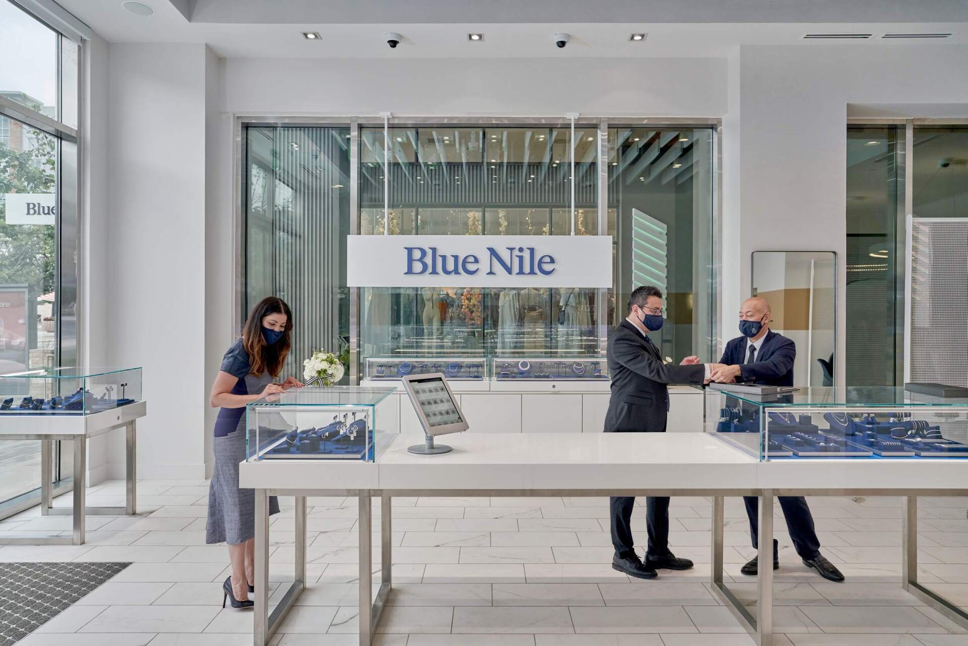 Blue Nile jewelry store exterior with staff interacting. Two men and a woman wearing masks behind display cases.