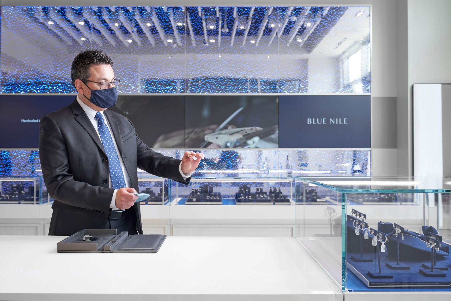 Man in suit and mask gestures in a luxury jewelry store with glass displays, blue decor.