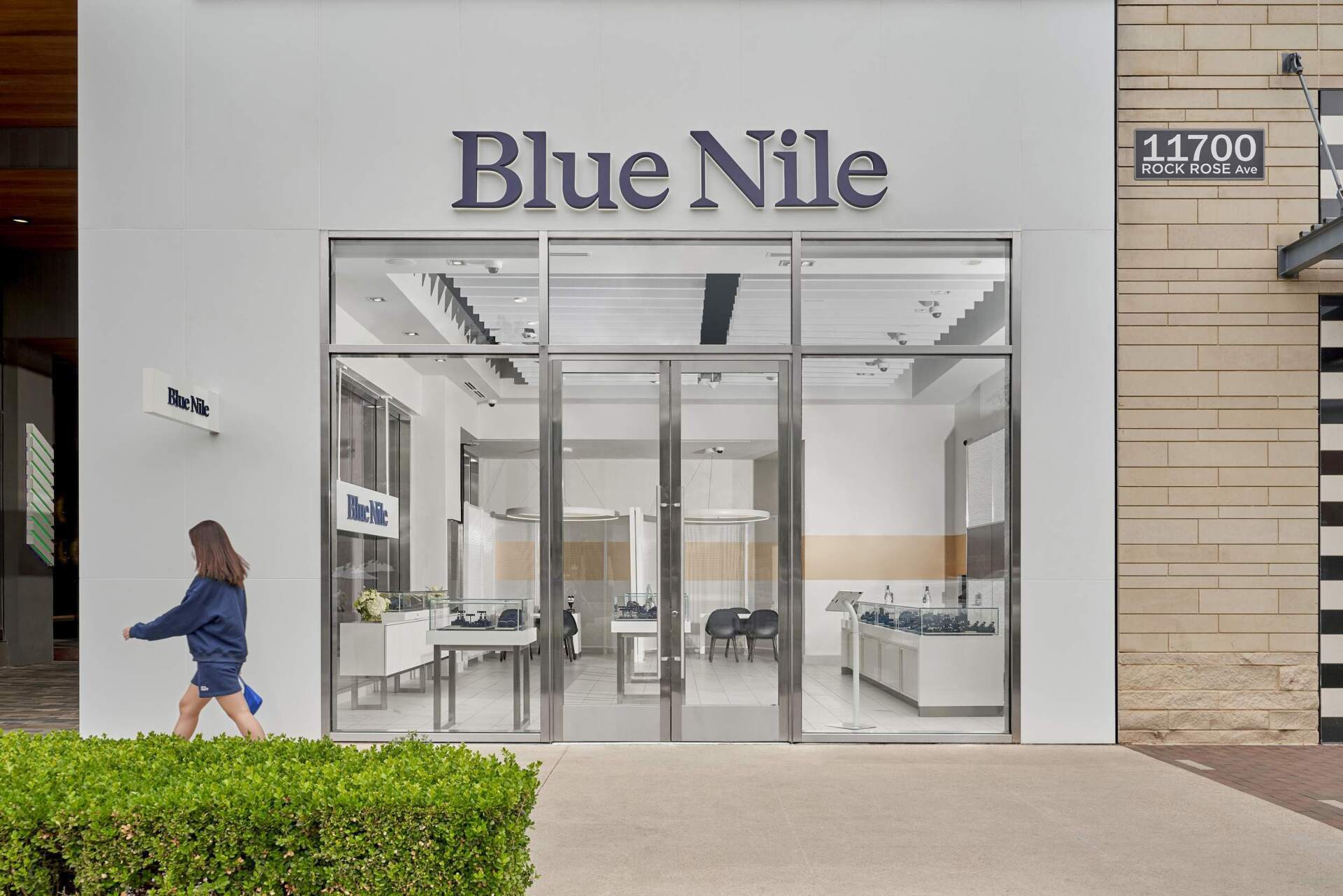 Exterior of a Blue Nile jewelry store with a pedestrian walking past. White building with large glass windows.
