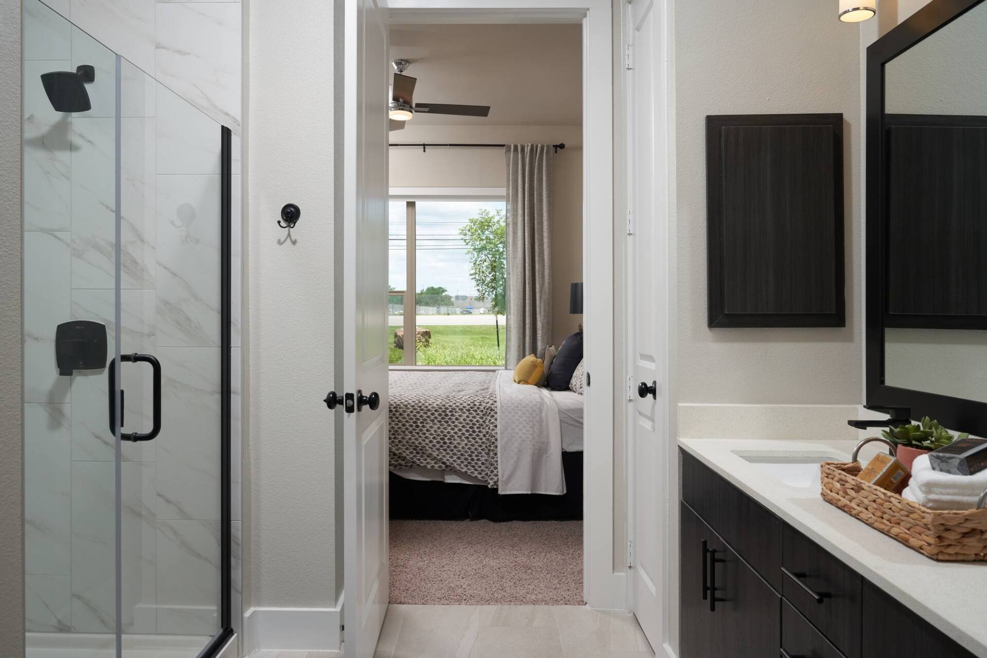 Bathroom doorway leading to a bedroom, featuring a shower, vanity, and a bed with a window view.