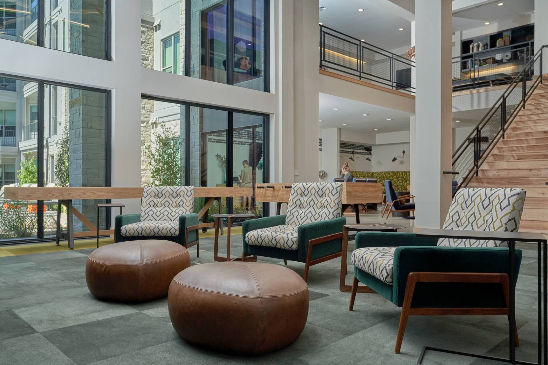 Lobby with seating: two ottomans, two armchairs with patterned cushions, large windows, wood accents, and staircase.