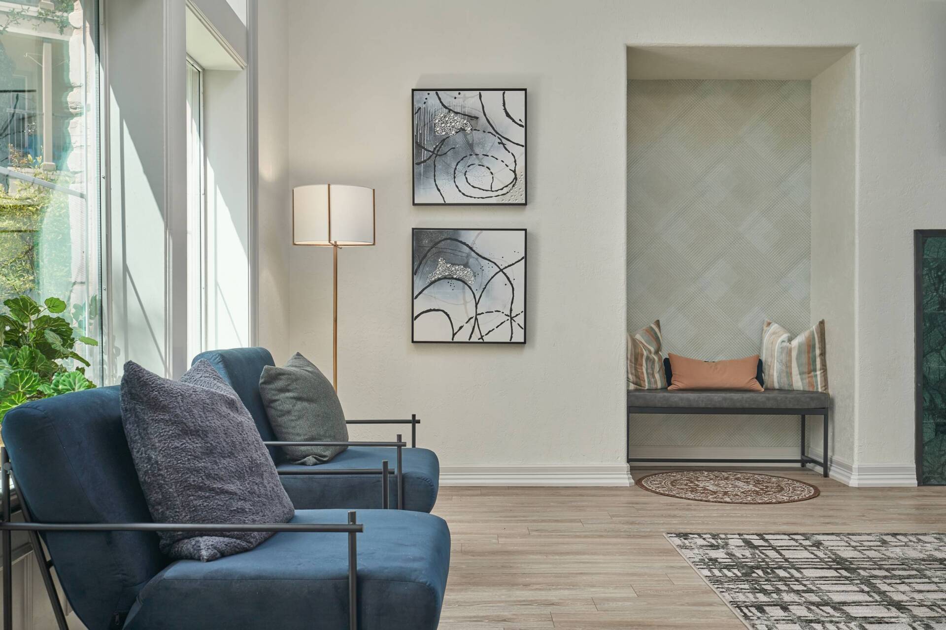 Two blue armchairs next to a floor lamp, art, and a bench in a neutral-toned room.