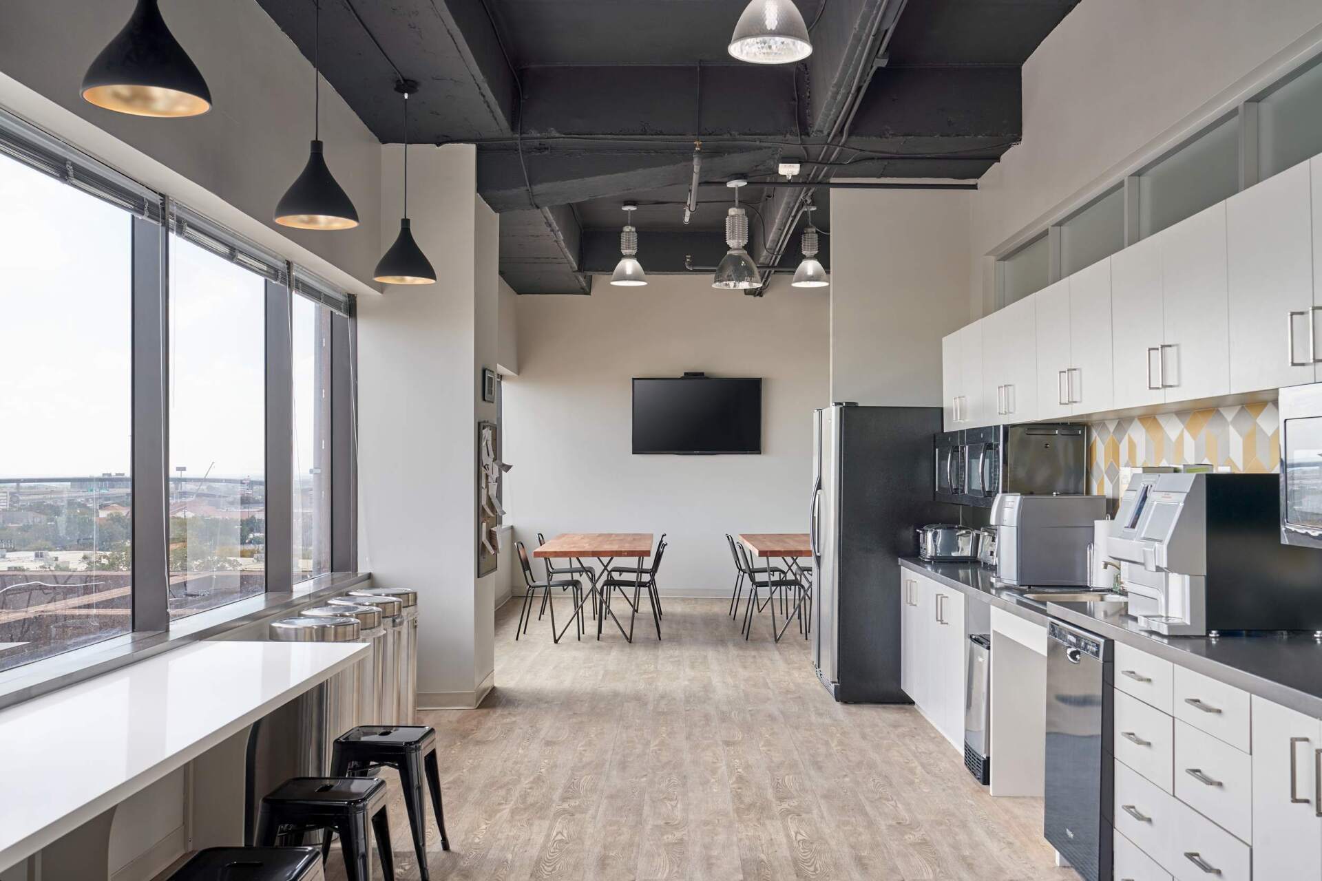 Office break room with large windows, tables, kitchenette, and black pendant lights.