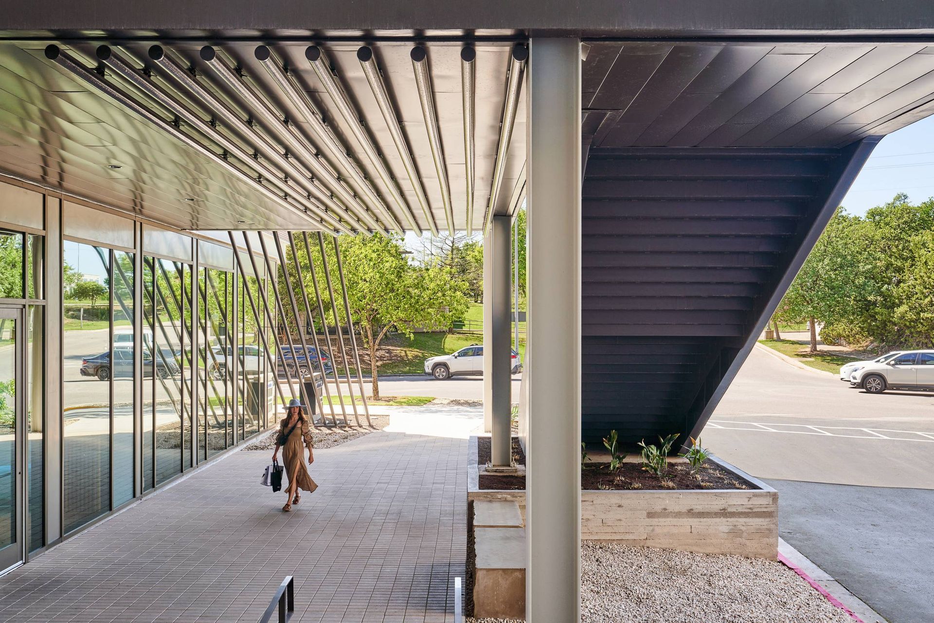 Woman walking under a covered walkway with glass wall; trees and cars in background.