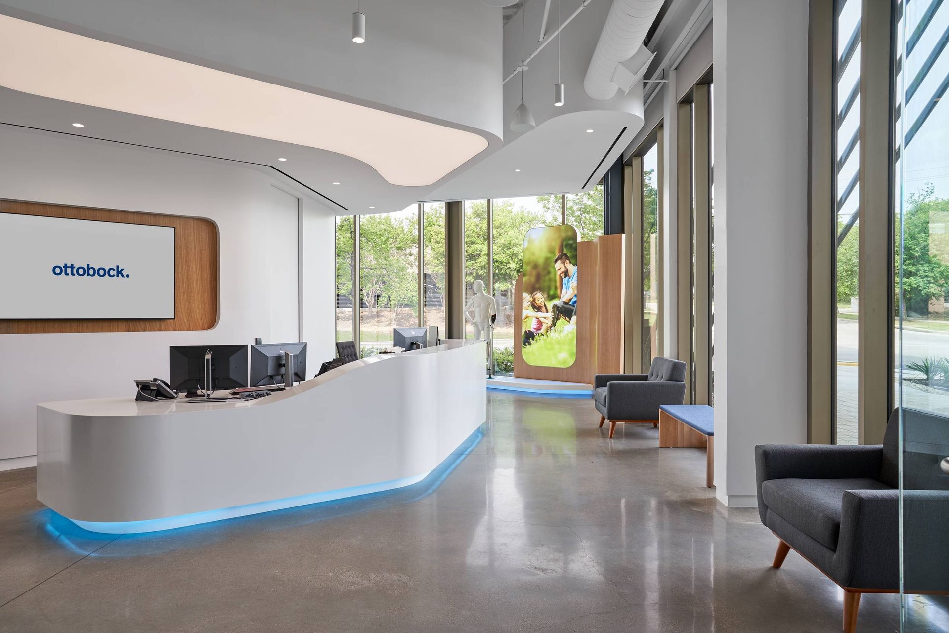 Modern office reception area with a curved white desk, a large screen, seating area, and large windows.