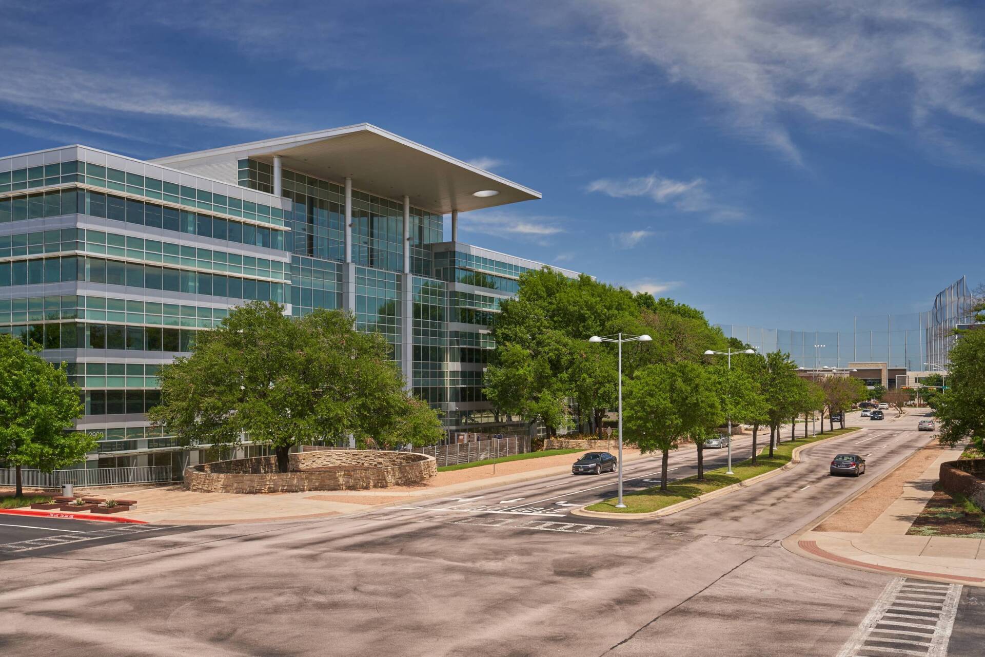 Modern multi-story office building with glass facade, trees, and street on a sunny day.