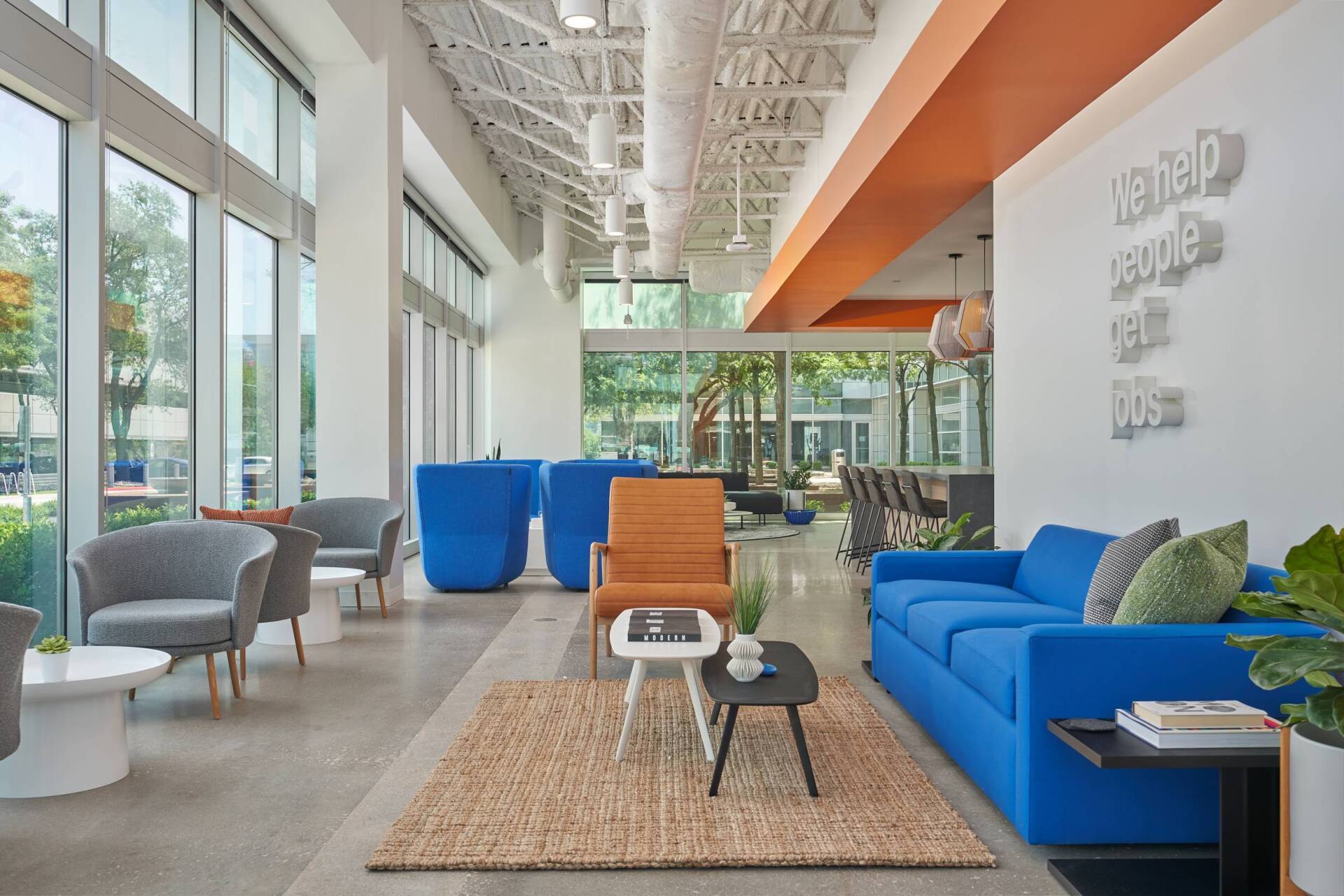 Modern office lobby with blue and orange accents, large windows, various seating, and white walls.
