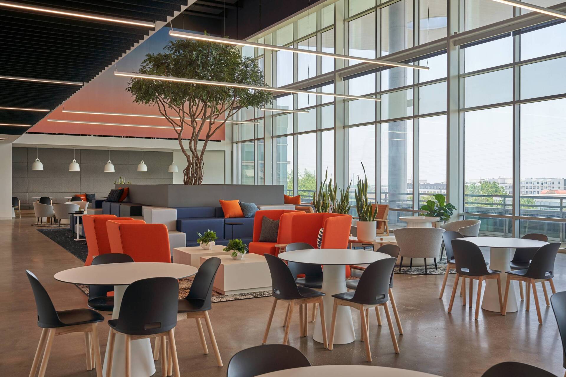 Modern office lounge with large windows, round tables, and colorful seating areas.