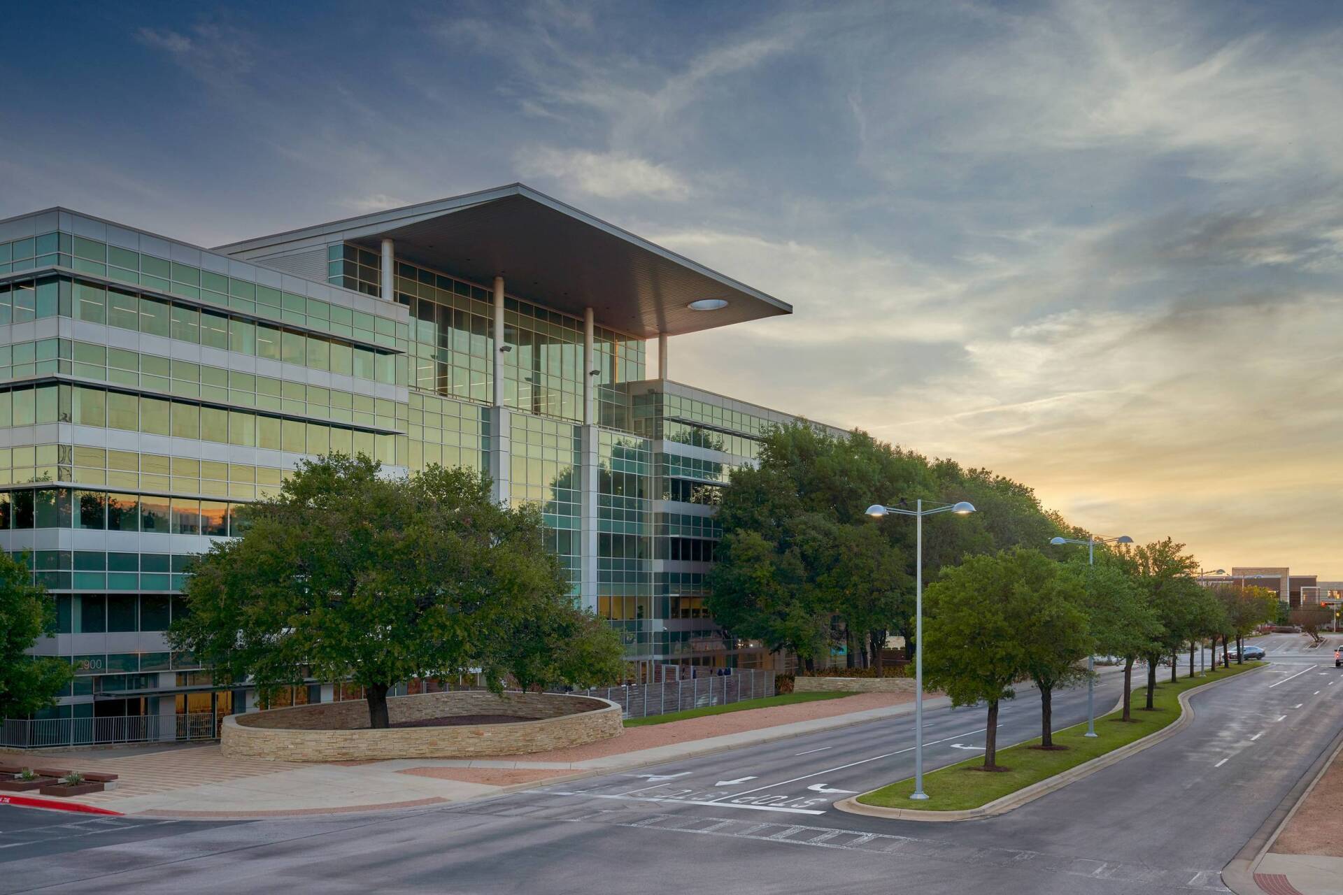 Modern glass office building with trees along the road and a cloudy sky.
