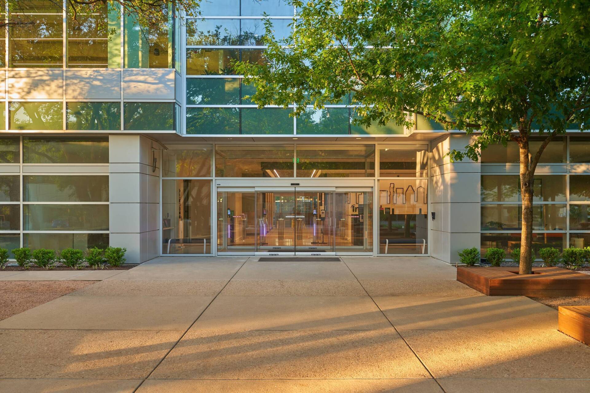 Modern building entrance with glass doors, surrounded by windows and trees. Sunlight casts shadows.