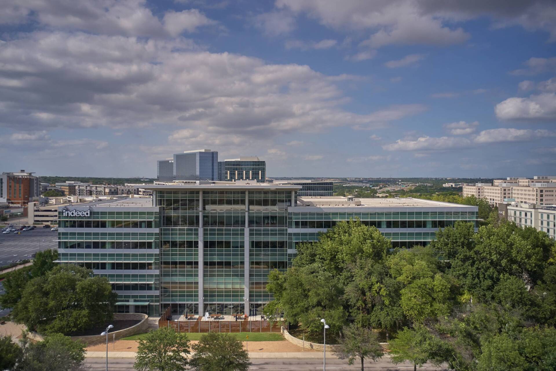 Modern office building with green glass facade, trees in front, and cloudy sky.