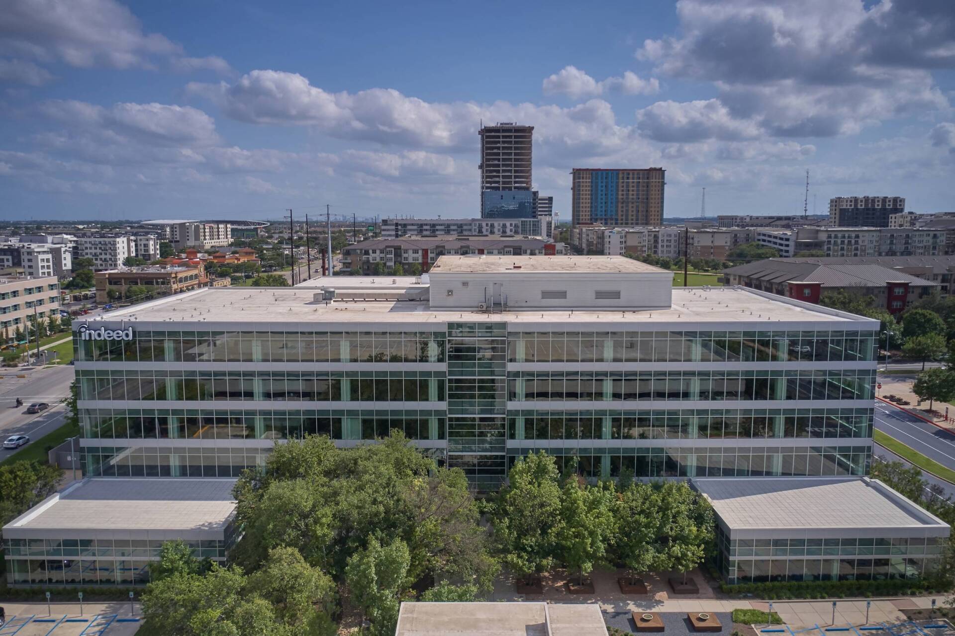 Modern glass-walled office building with trees in front, set in an urban environment under a blue sky.