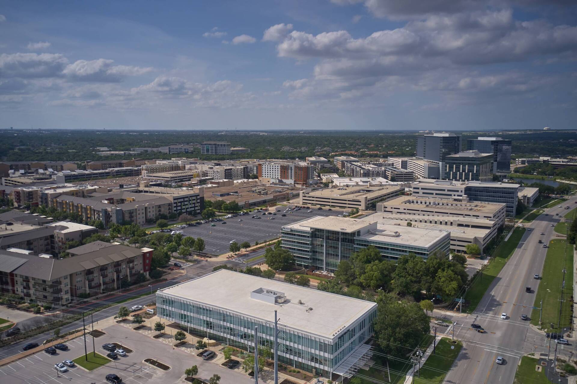 Aerial view of a city with buildings, parking lots, and roads on a sunny day with clouds.