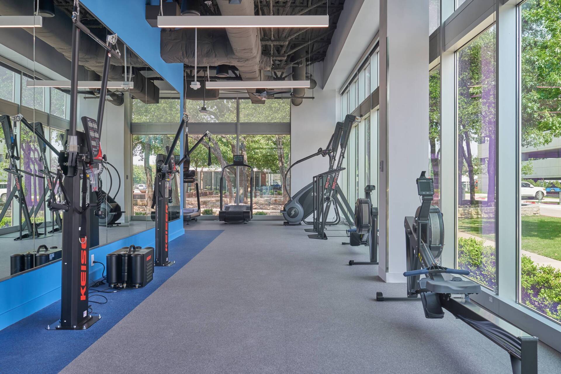 Gym interior with exercise equipment near windows. Gray carpet, blue accents, natural light.
