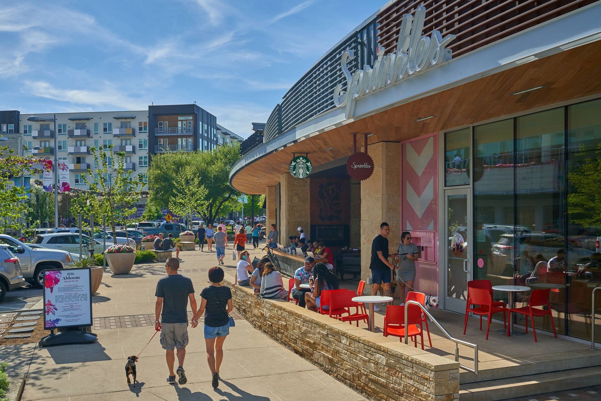 People walking on a sunny sidewalk in front of a Starbucks with outdoor seating.