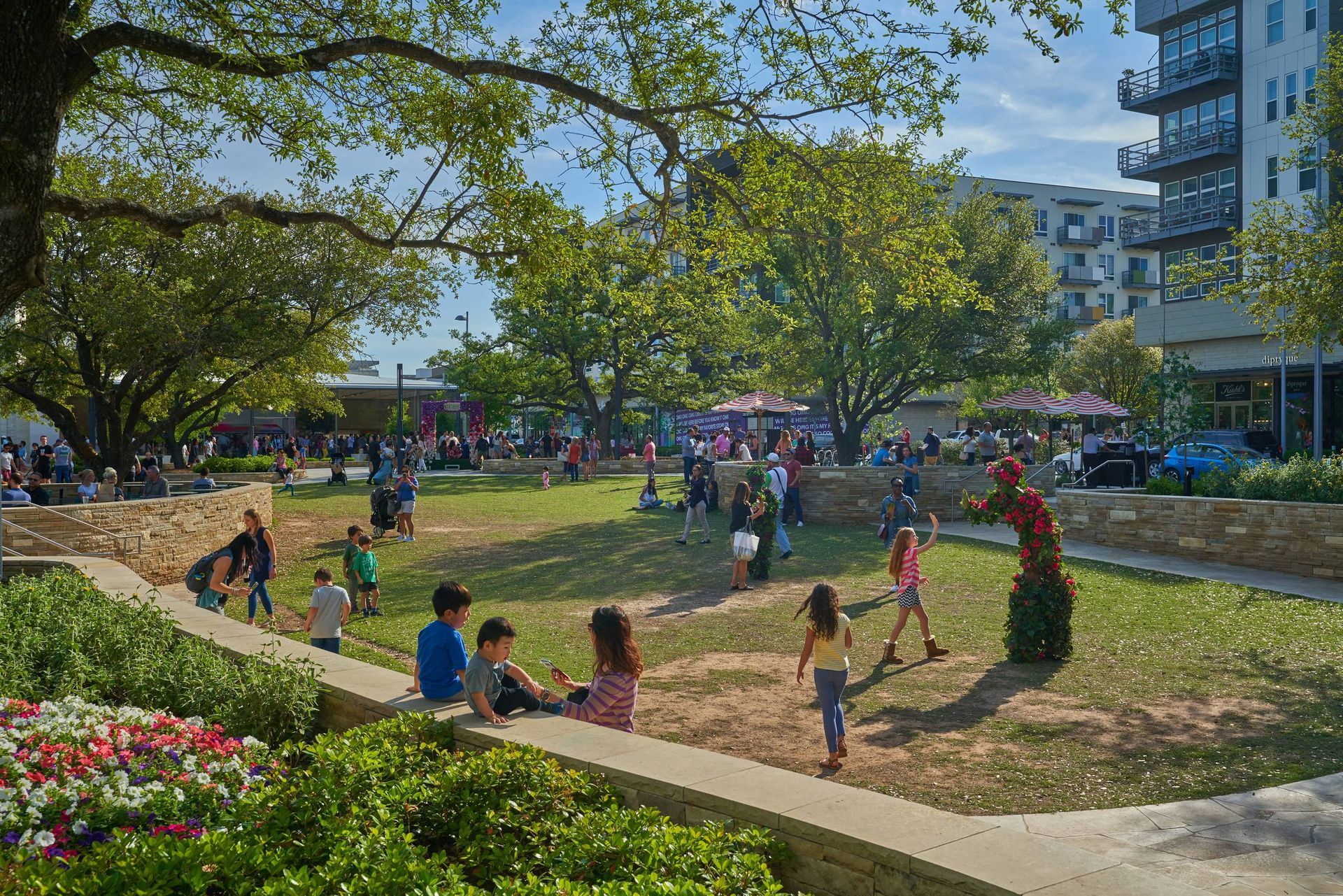 Park with people, trees, and buildings. Children play on grass, others sit. Sunny day.