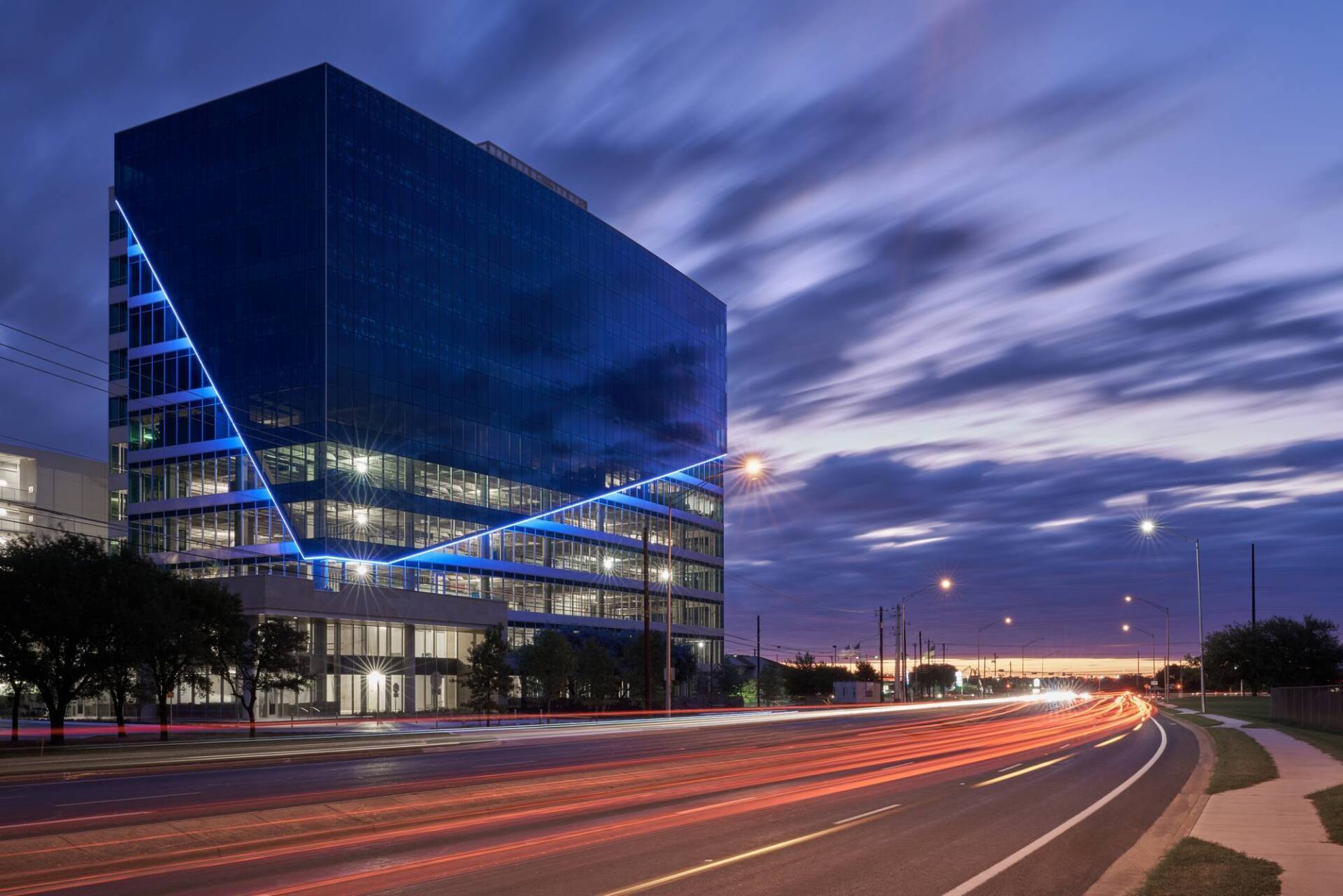 Glass office building with blue accents at dusk, streaks of light from passing cars.