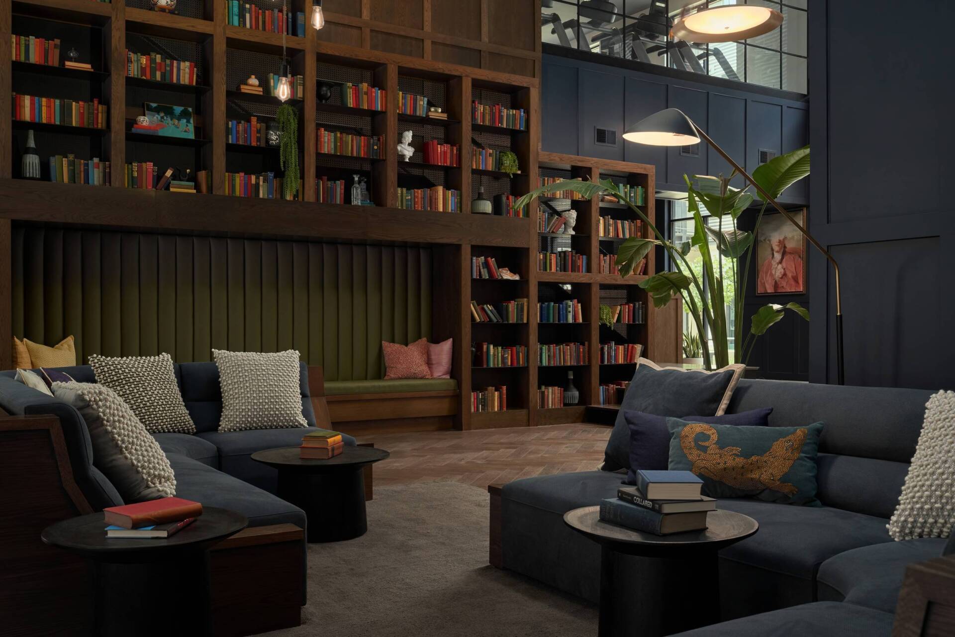 Cozy library lounge with dark blue couches, bookshelves, small tables. Green and brown tones.