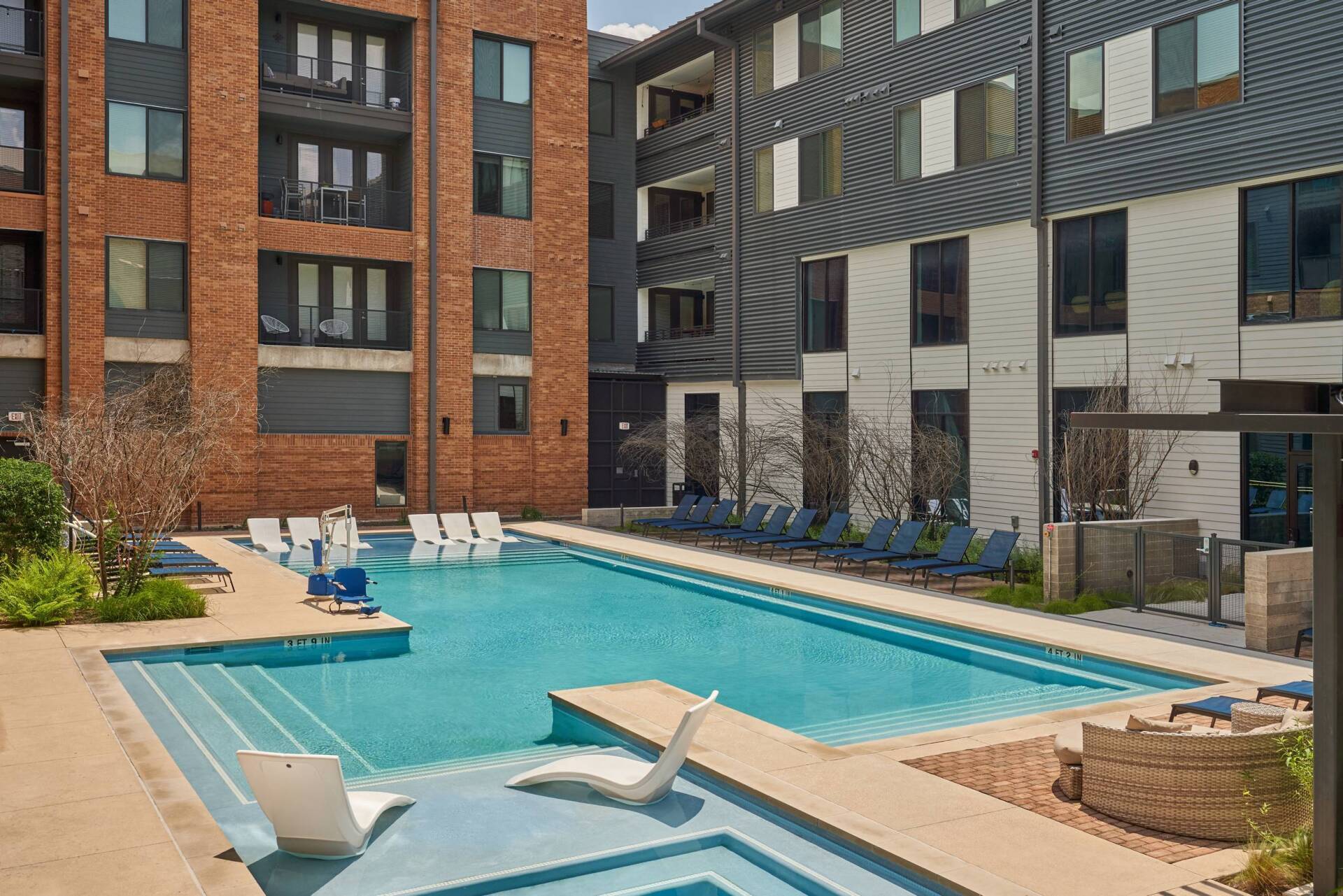Swimming pool in a courtyard surrounded by modern apartment buildings; lounge chairs and trees.