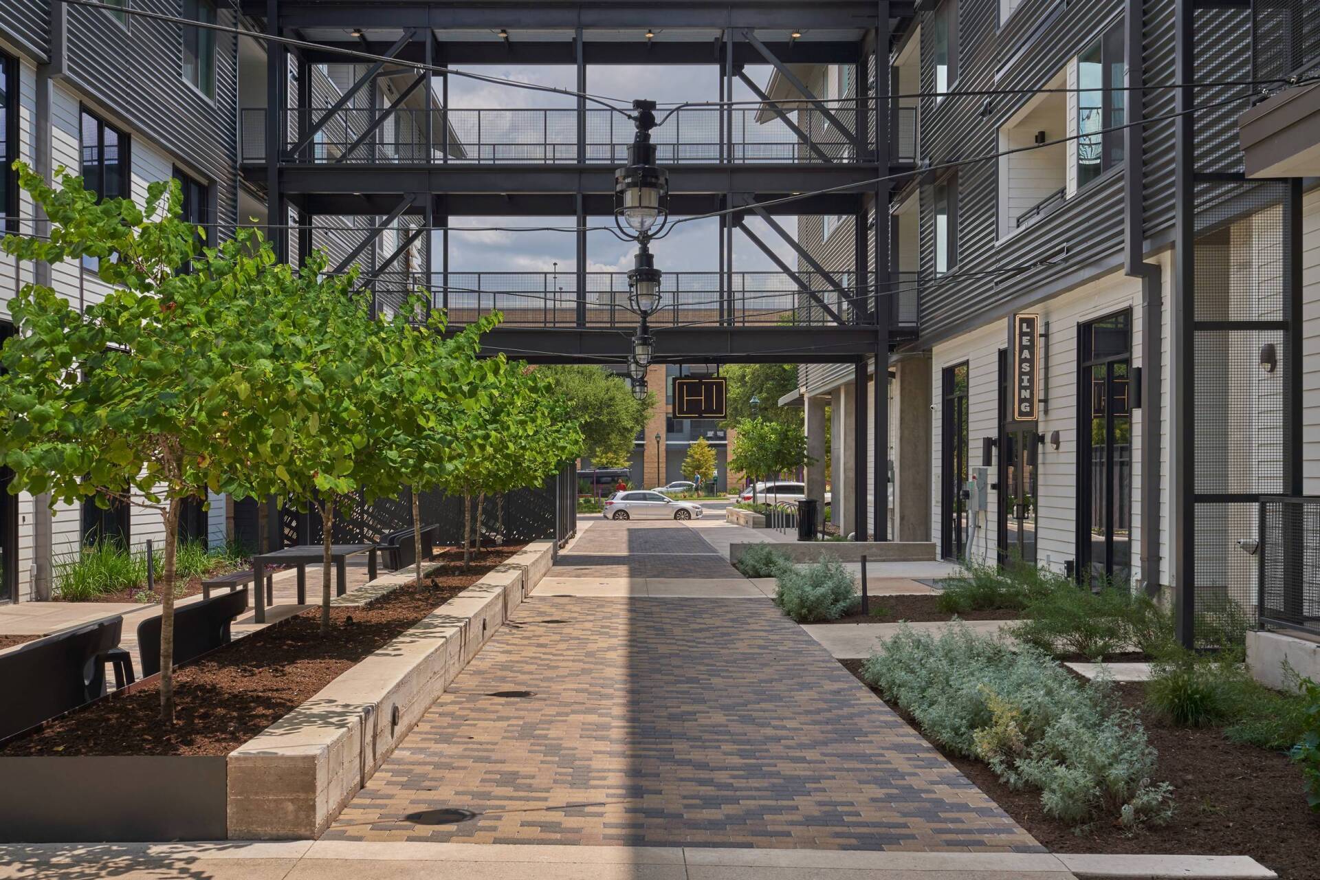 Courtyard with brick path, trees, seating, and a black metal bridge structure overhead.