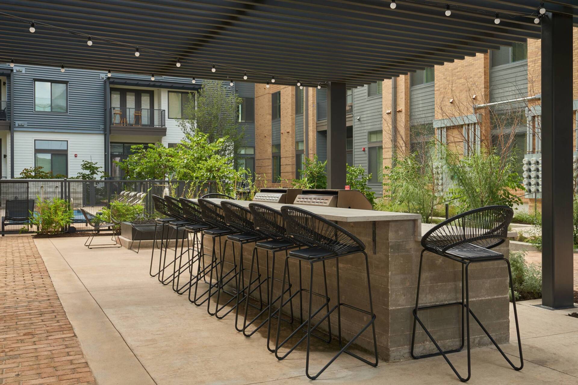 Outdoor kitchen with built-in grill and bar seating under a pergola. Brick patio, modern buildings in background.