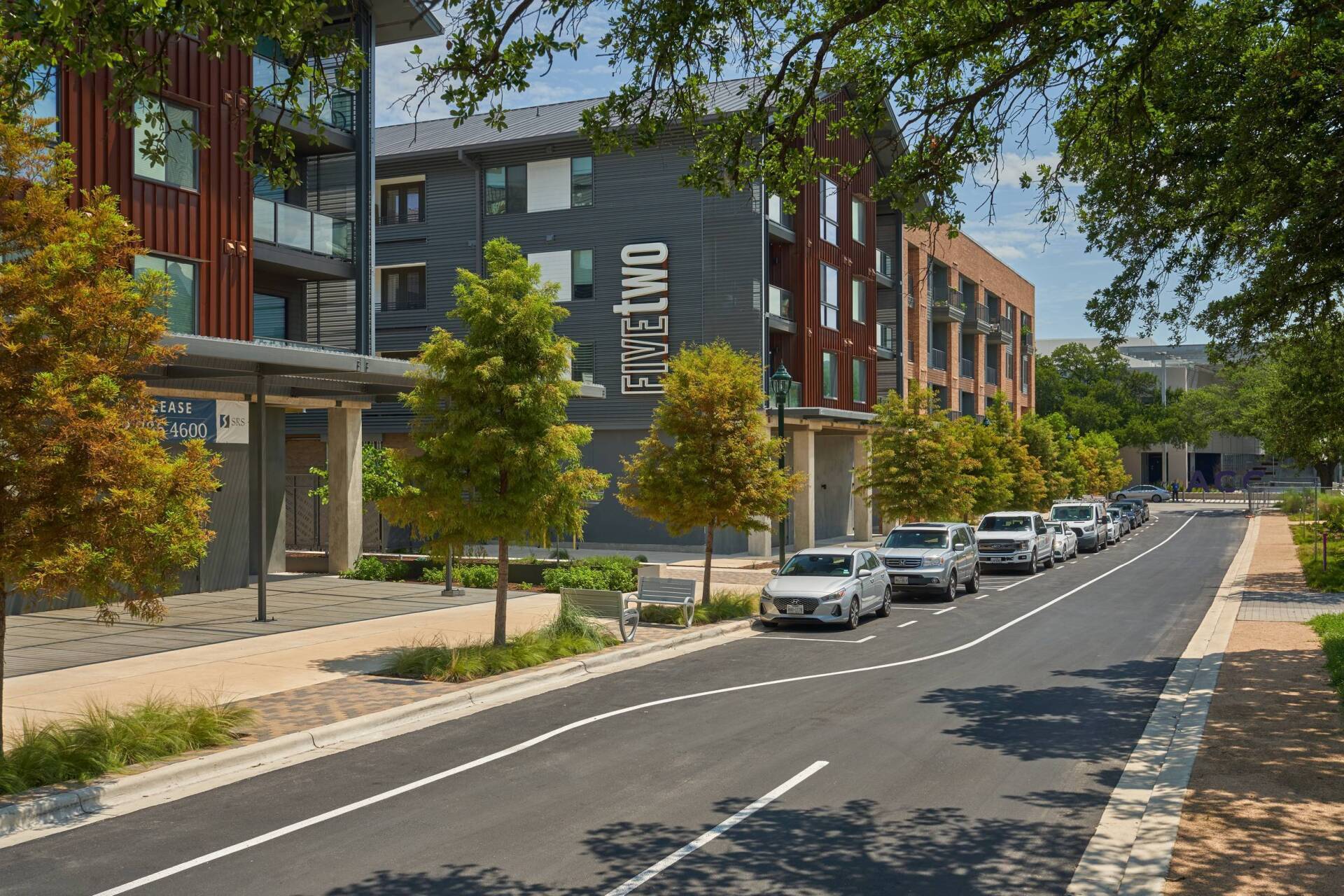 Street lined with parked cars and trees in front of modern apartments with “Twelve” on the building.
