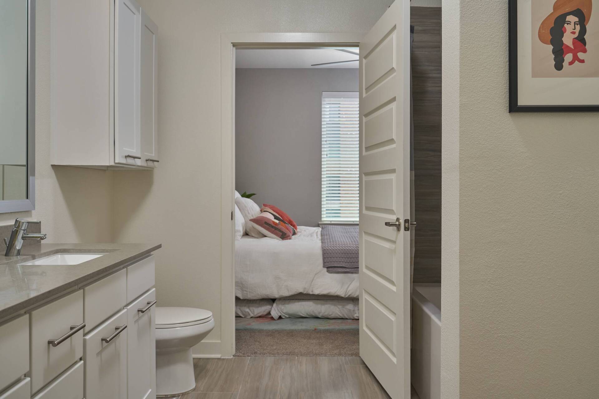 Bathroom doorway leading to a bedroom with bed and window blinds. White cabinets and grey countertop.