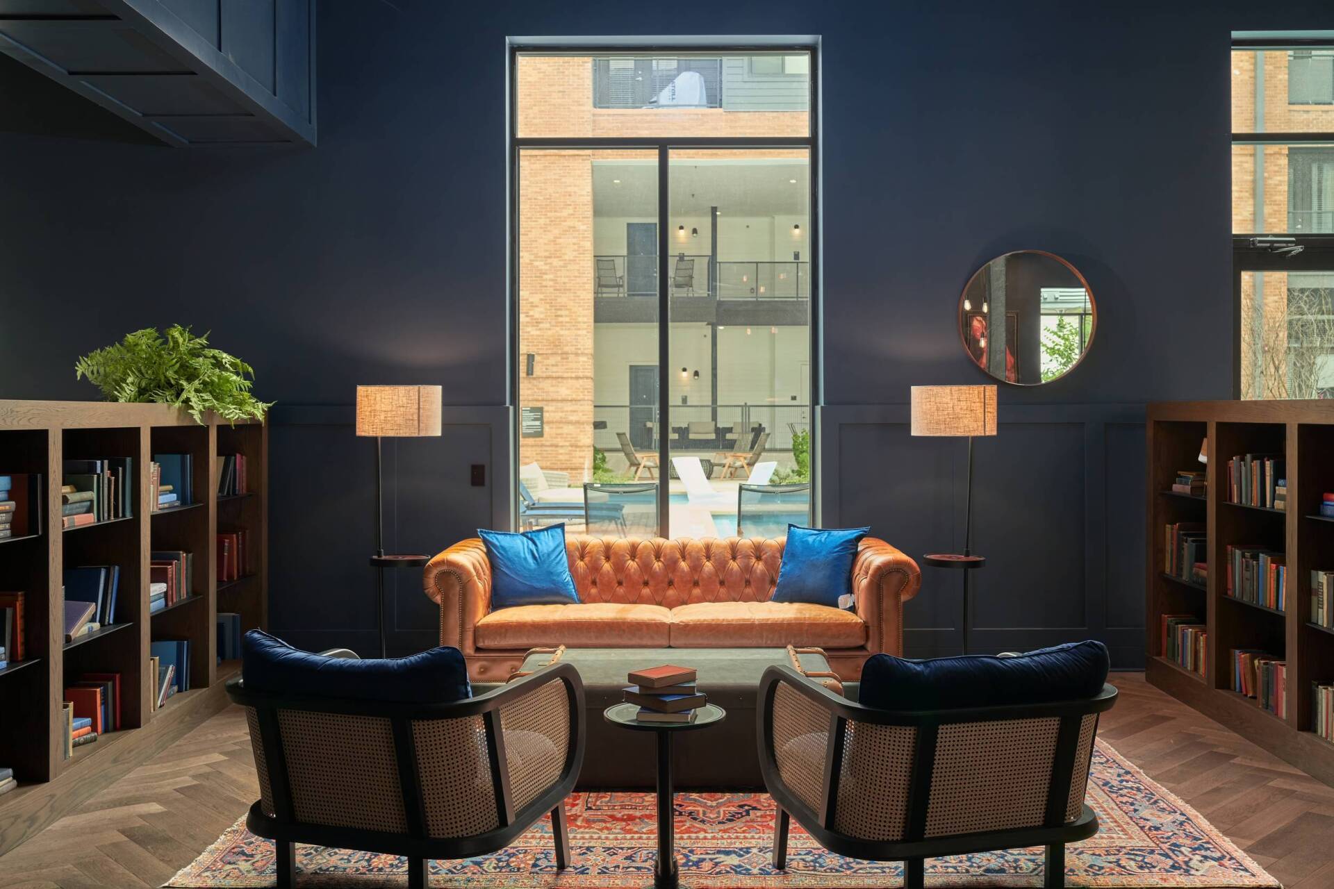 Library with blue walls, tan leather sofa, two chairs, and floor lamps by a large window overlooking a courtyard.