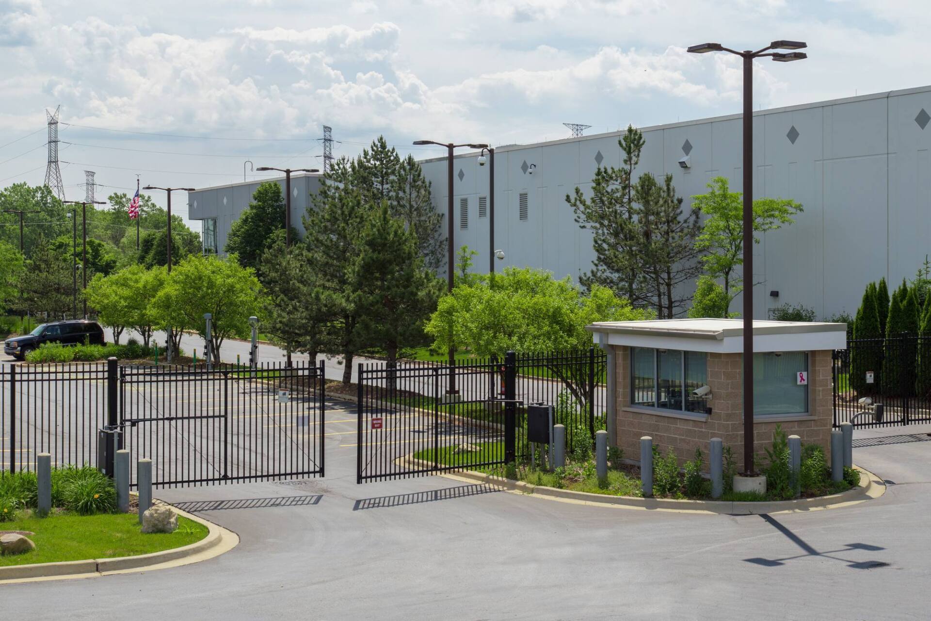 Gated entrance to a large industrial building, with a security guard booth and trees.