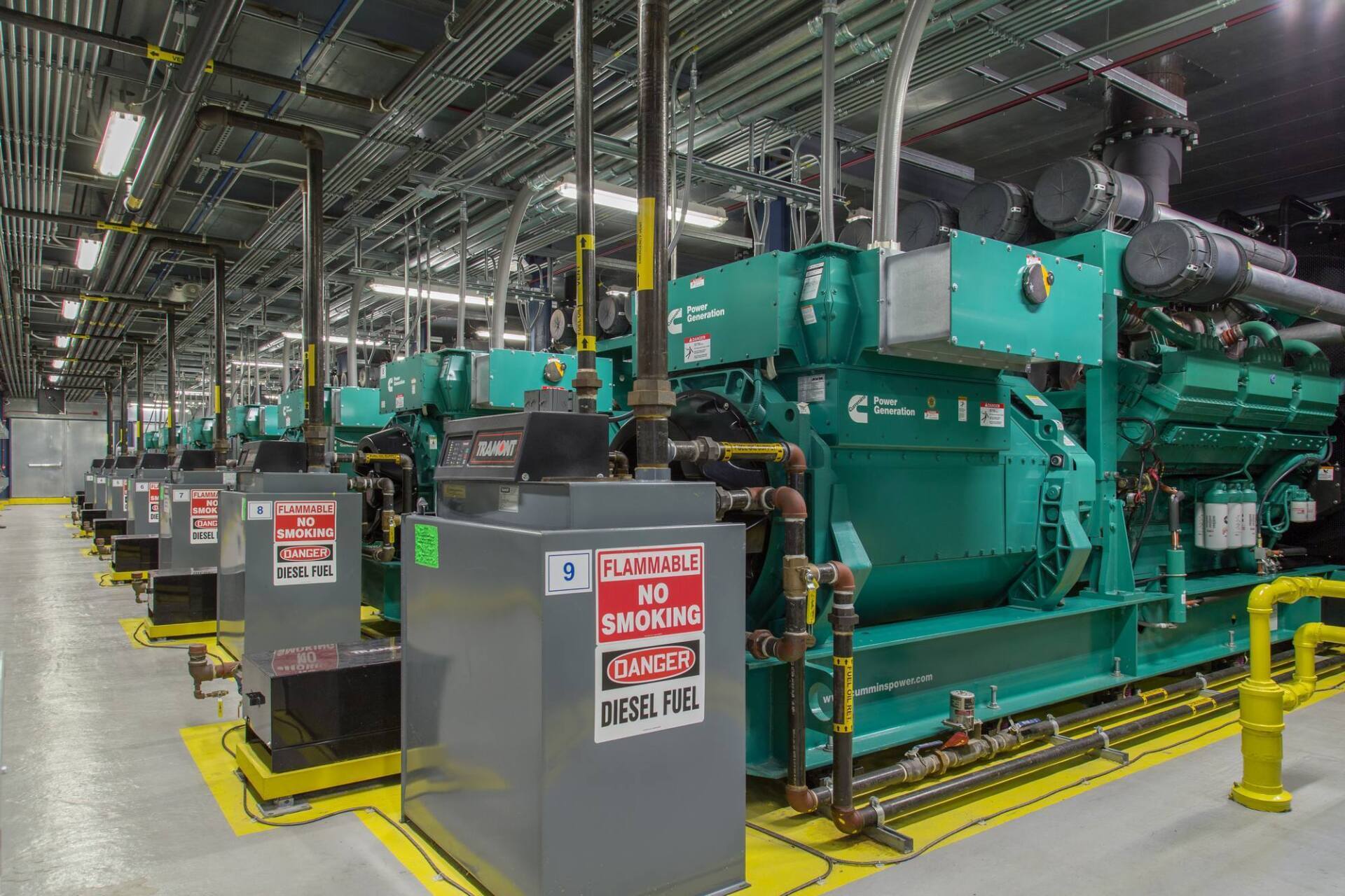 Green industrial diesel generators in a utility room, with gray control boxes and warning labels.
