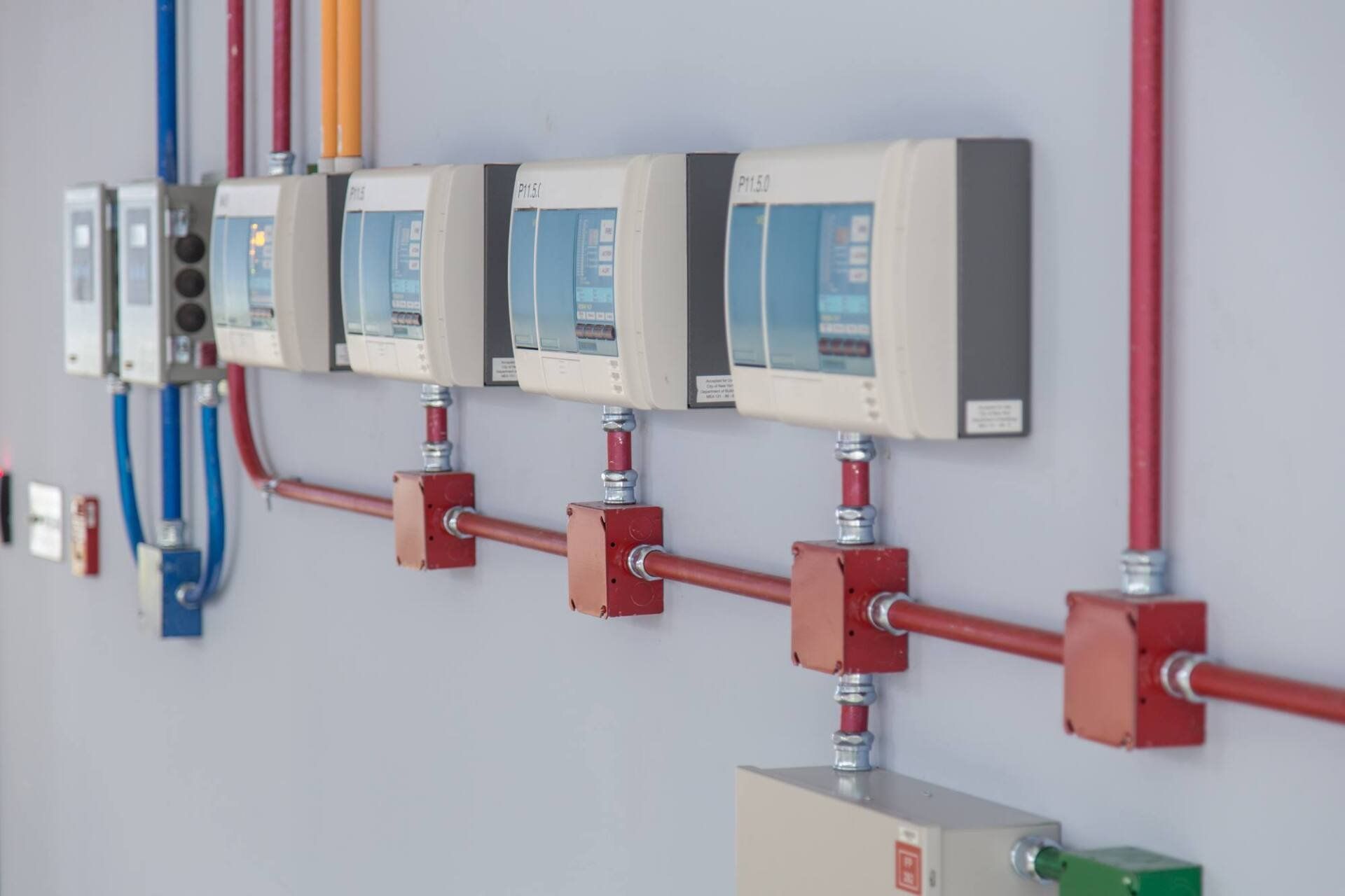 Electrical conduit and junction boxes mounted on a white wall, with red, blue, and yellow pipes.
