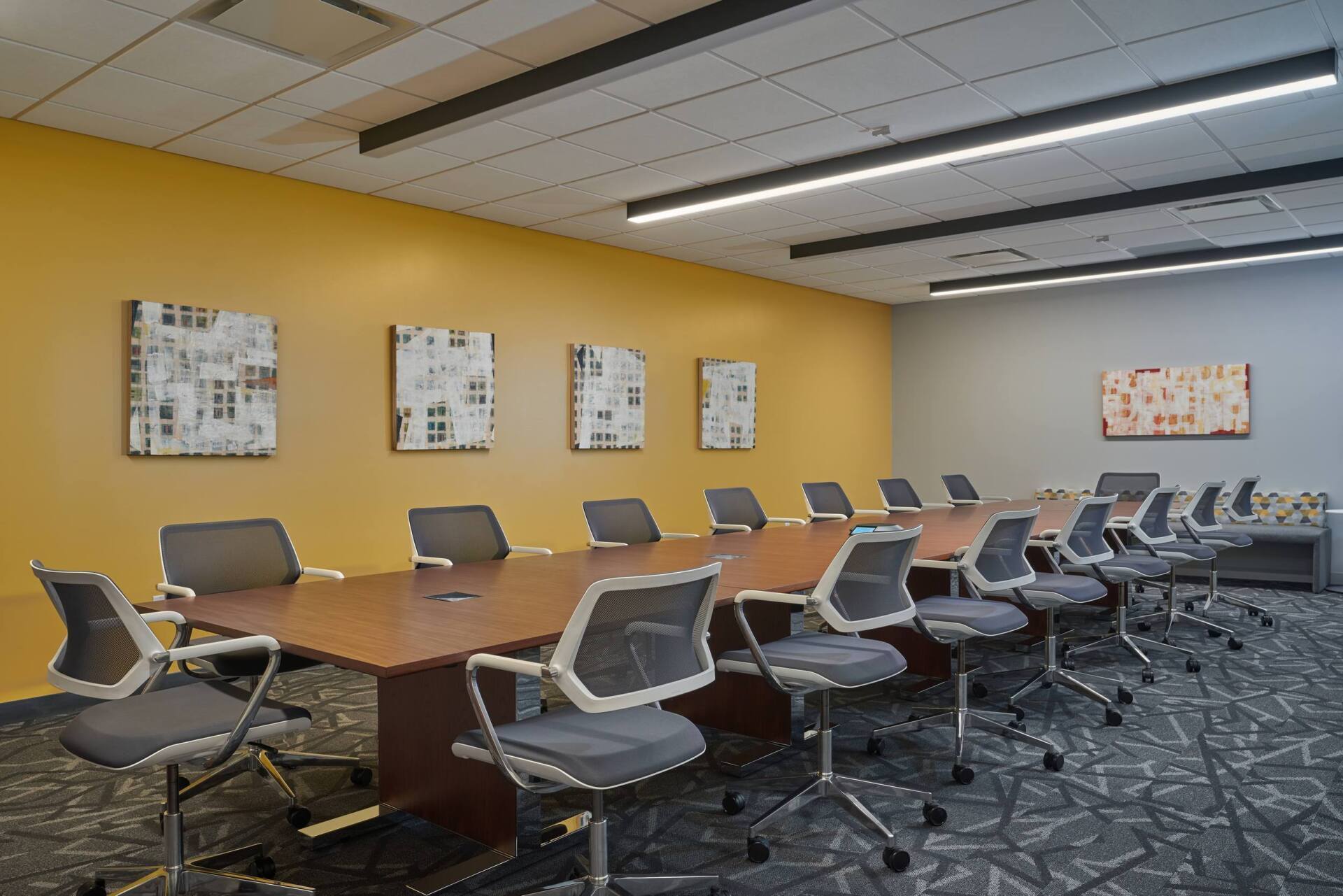 Conference room with long wooden table, gray chairs, yellow and gray walls, art, and patterned carpet.