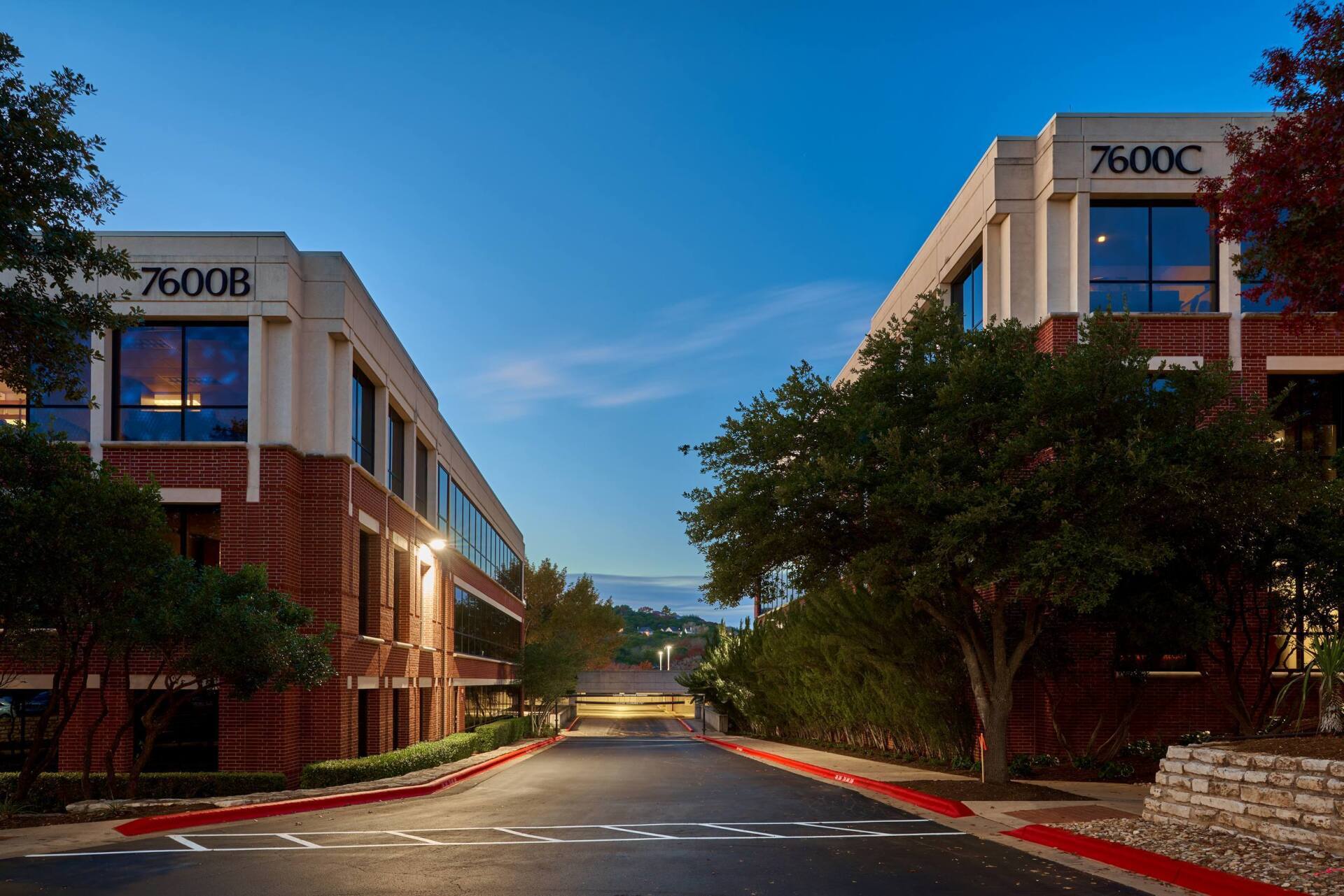 Two office buildings with brick exteriors and address signs, a paved road, and trees.