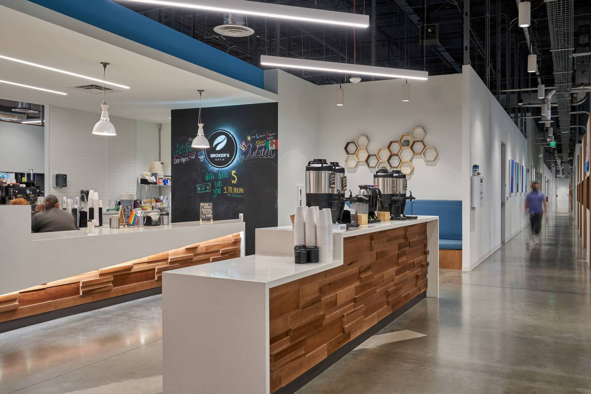 Modern office break room with coffee station, white counters, wood paneling, and a person walking in the hallway.