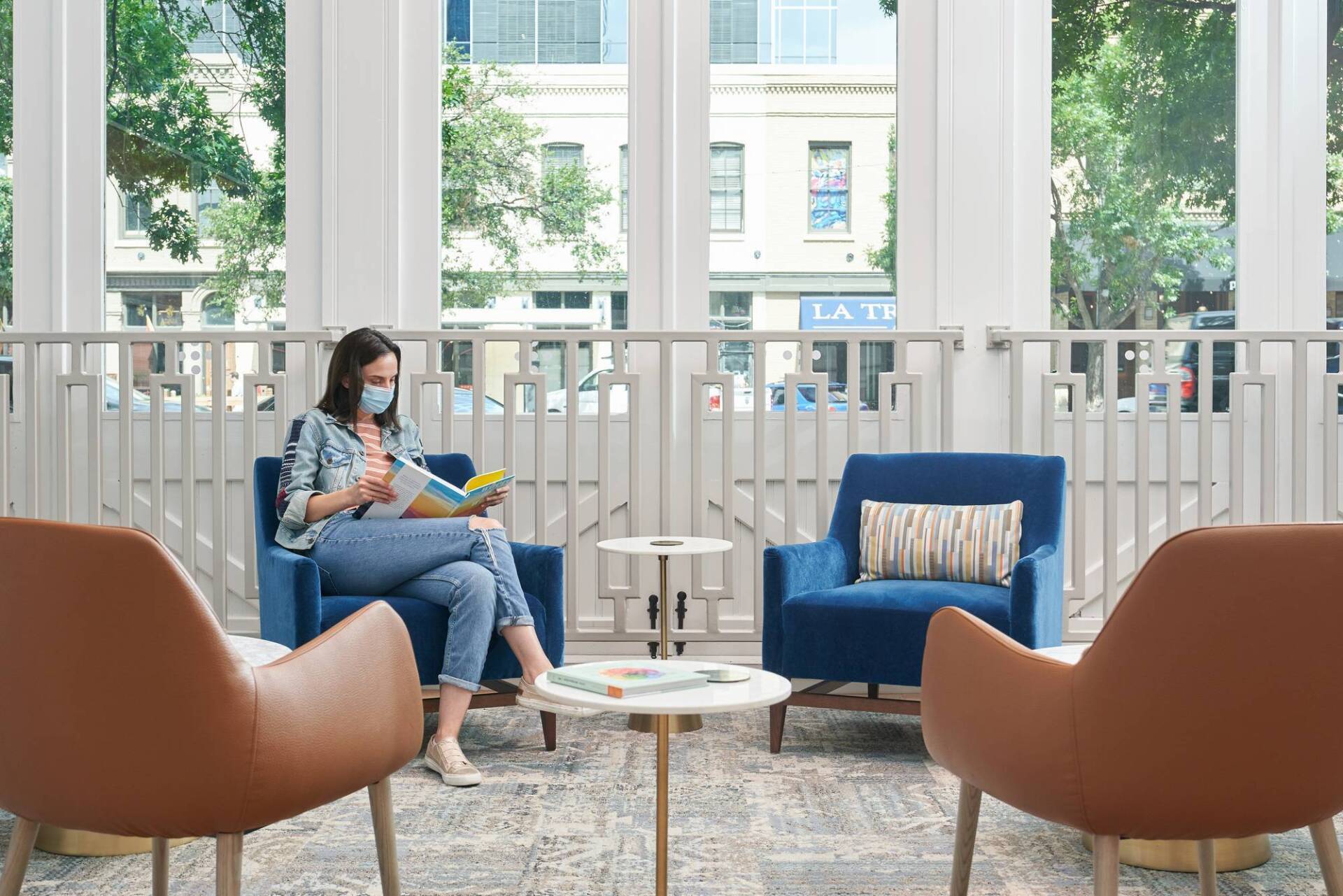 Woman in mask reads in a blue armchair near a window. Two other chairs face the scene, city view.