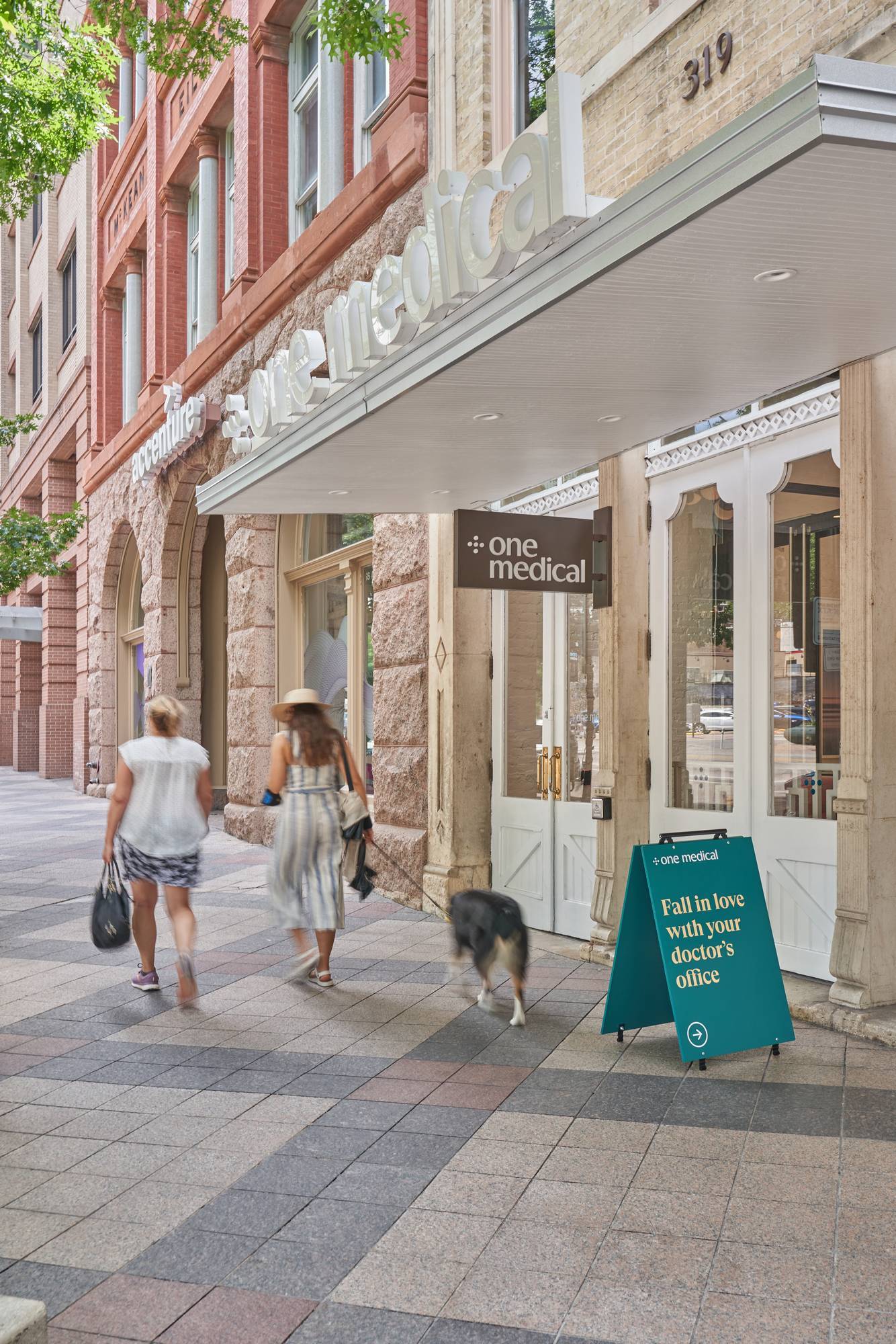 People walking on a sidewalk toward a storefront with a dog, under a white awning.